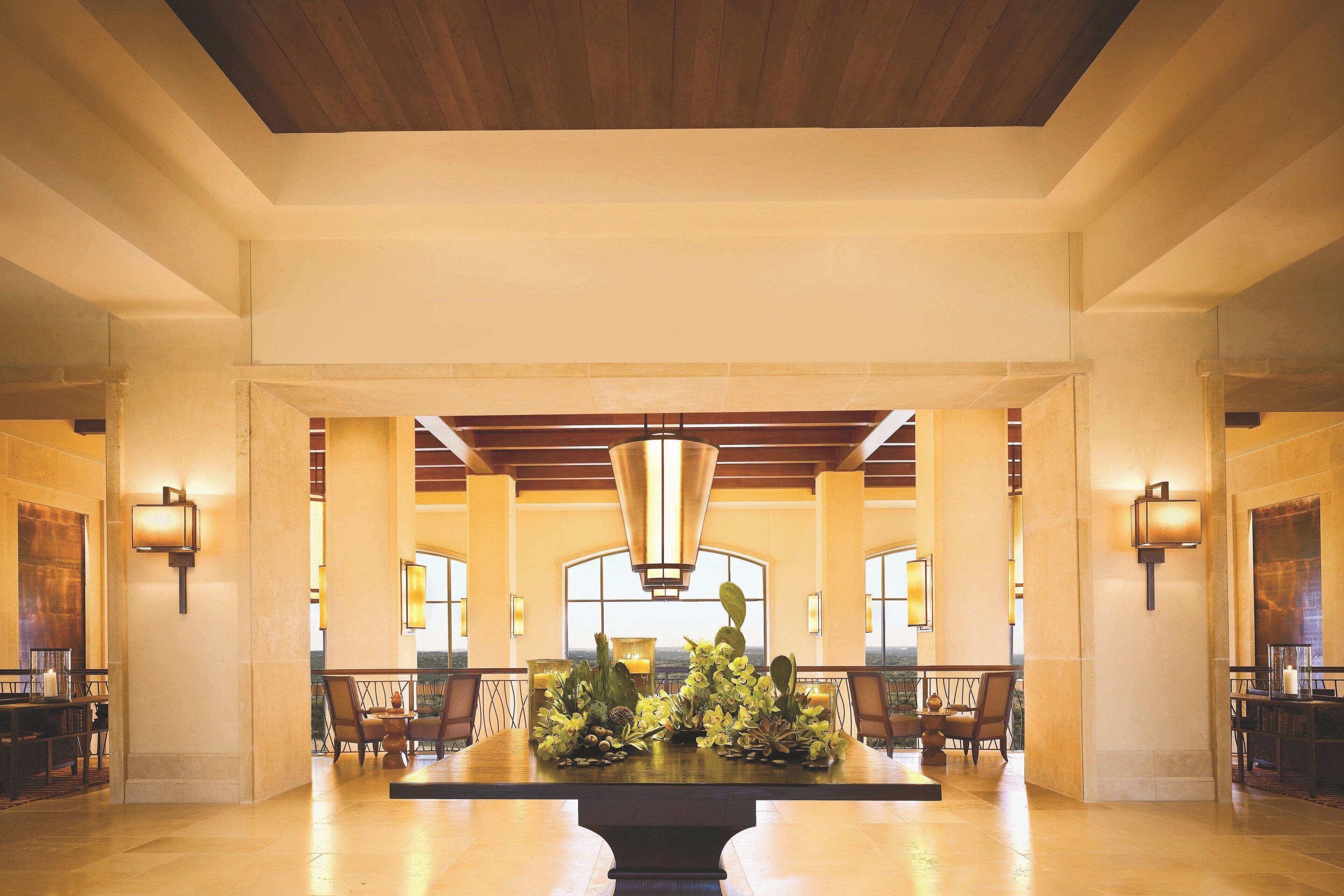 A spacious lobby with a central table adorned with flowers, high ceilings with wooden beams, and large windows providing natural light.