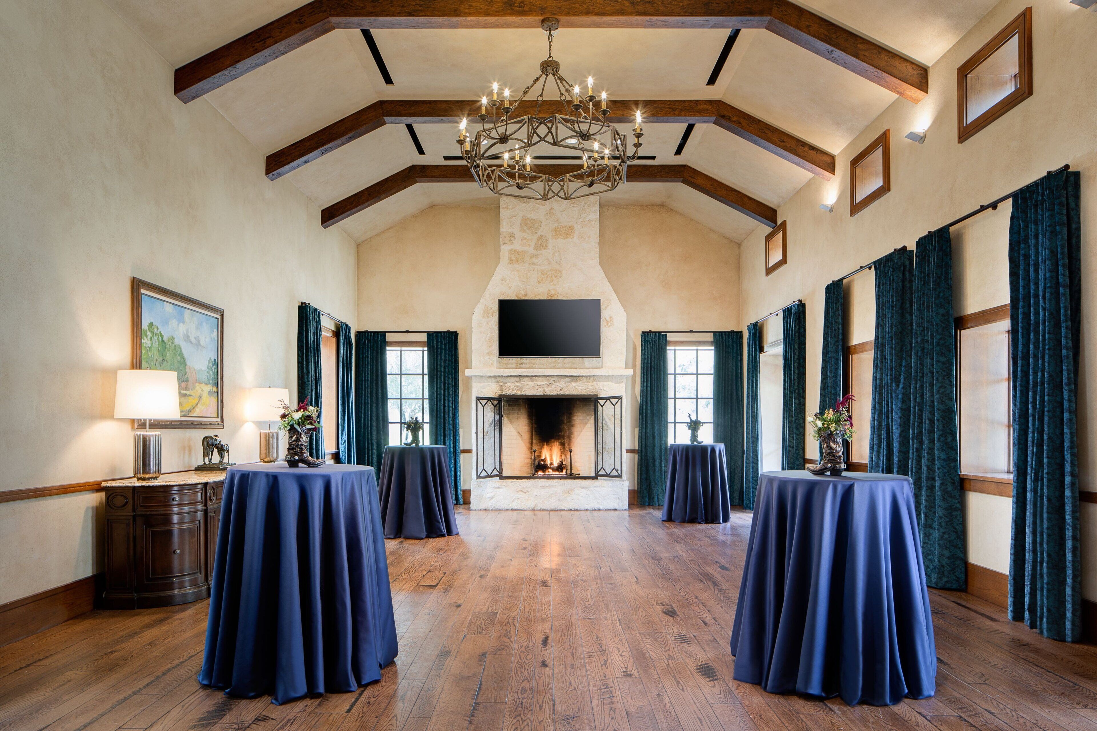 A spacious room with a fireplace, wooden beams, and a chandelier. Tables are set up for dining.