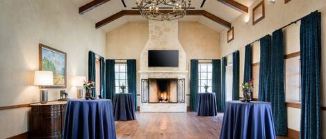 A spacious room with a fireplace, wooden beams, and a chandelier. Tables are set up for dining.