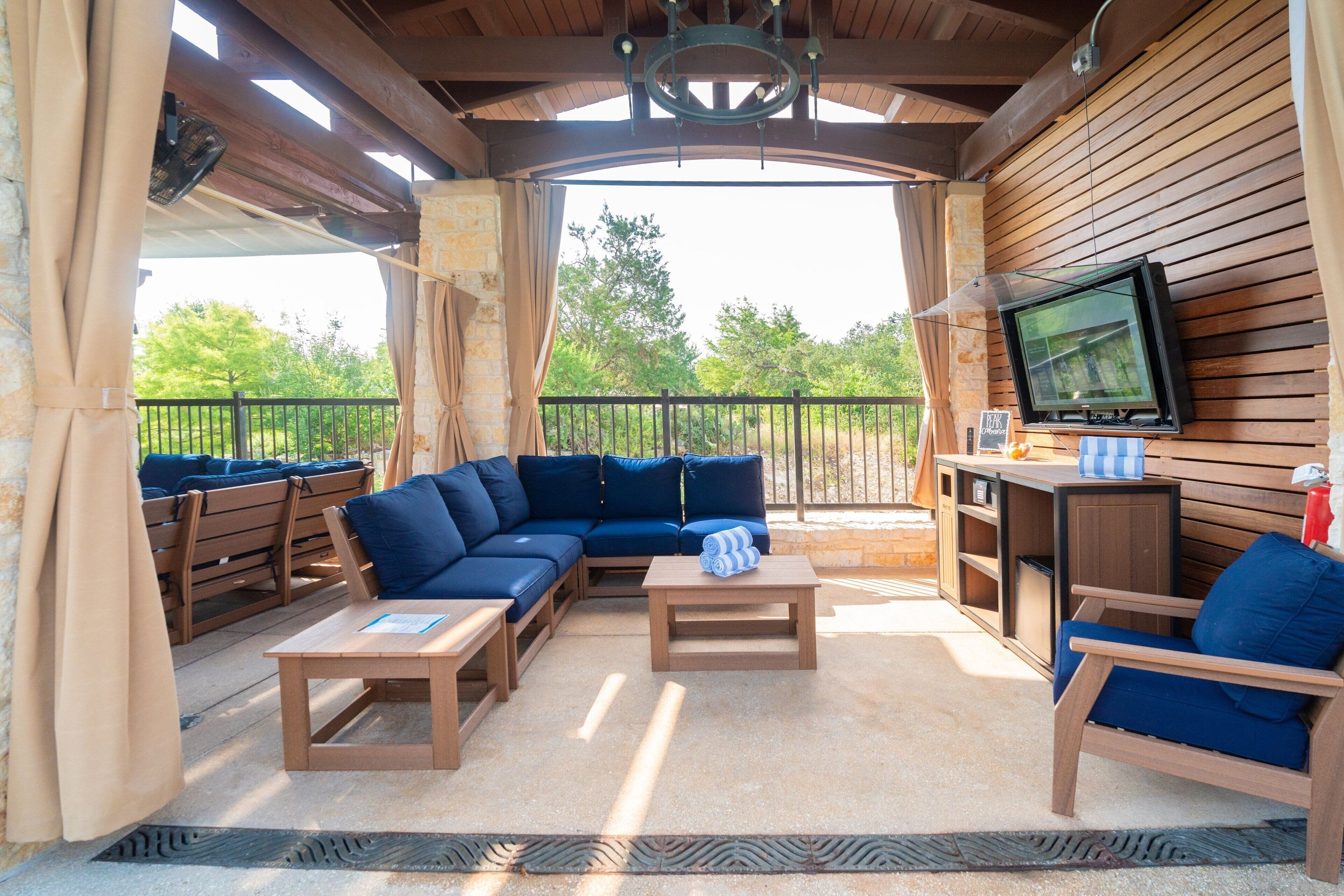 An outdoor patio with a wooden ceiling, a sectional sofa, a TV mounted on the wall, and a small table with a cushion.