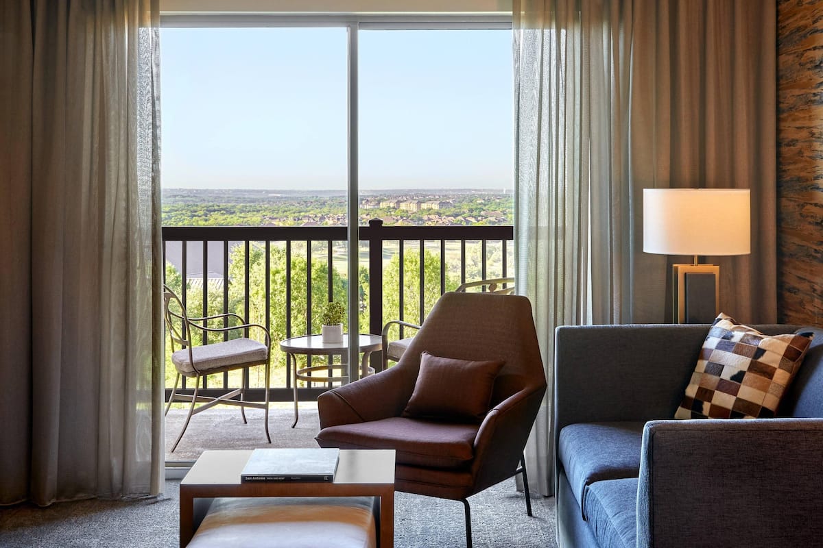 Una habitación de hotel con balcón, sofá, sillón, mesita y lámpara.