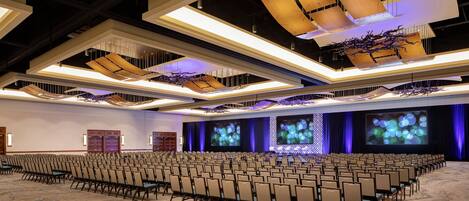 Un amplio salón de conferencias con filas de sillas, un escenario con una pantalla grande y luminarias modernas.