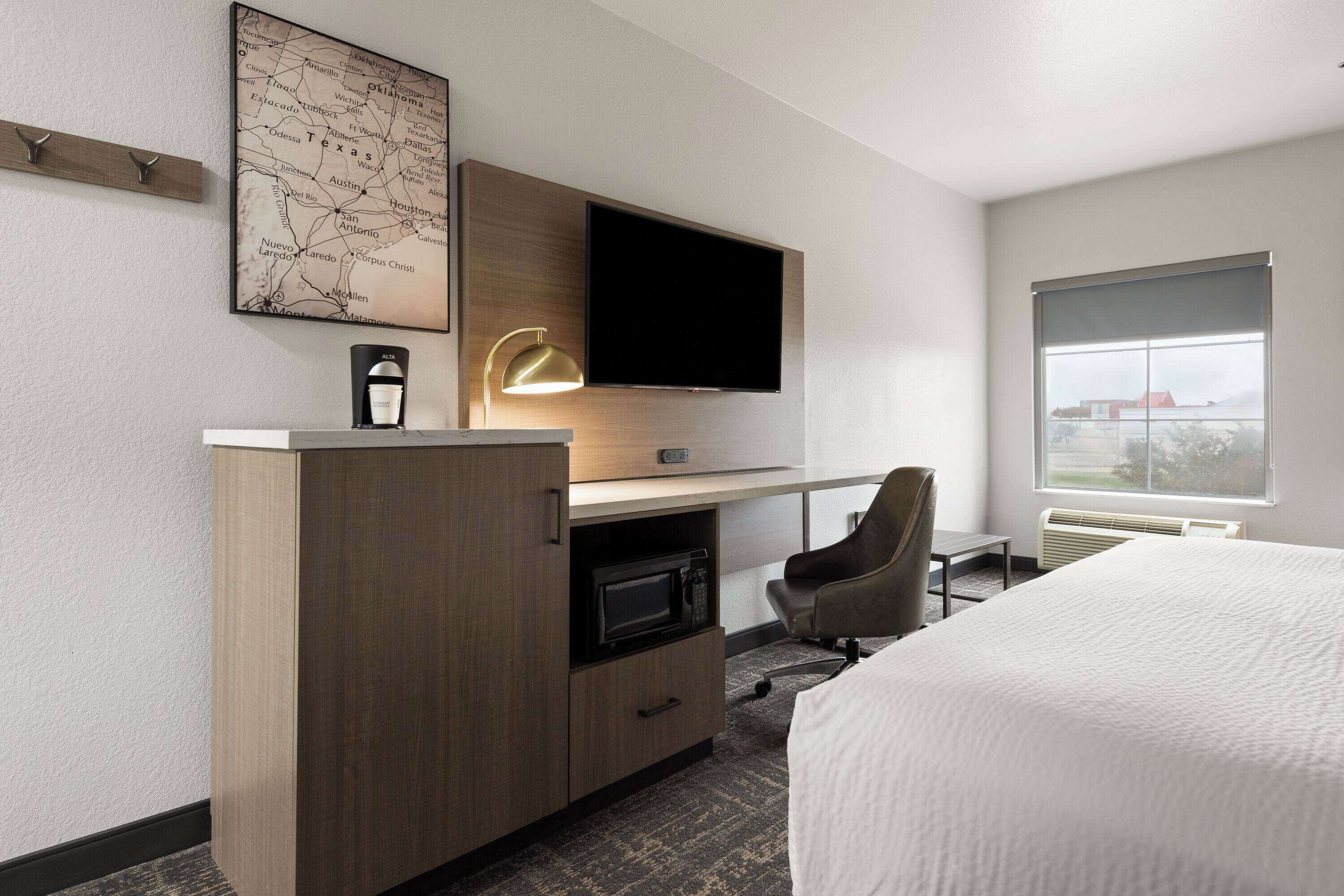 A hotel room with a bed, a desk with a flat-screen TV, a chair, a microwave, a map on the wall, and a window with a view.