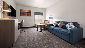 Una habitación de hotel moderna con un sofá azul, una mesita, una silla, un escritorio con lámpara, un televisor de pantalla plana y un cuadro en la pared.