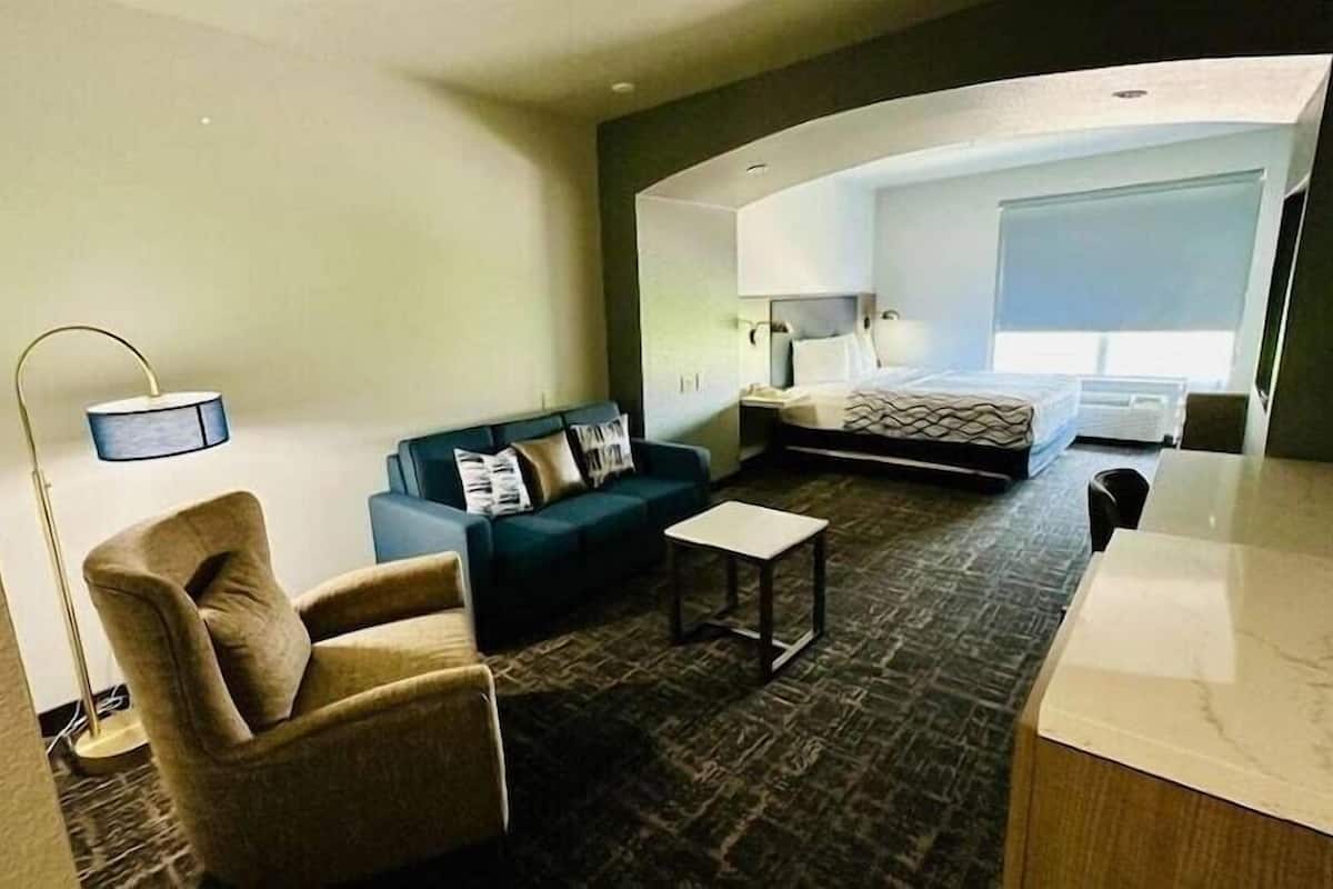 A hotel room with a sofa, armchair, small table, and a bed with a headboard.