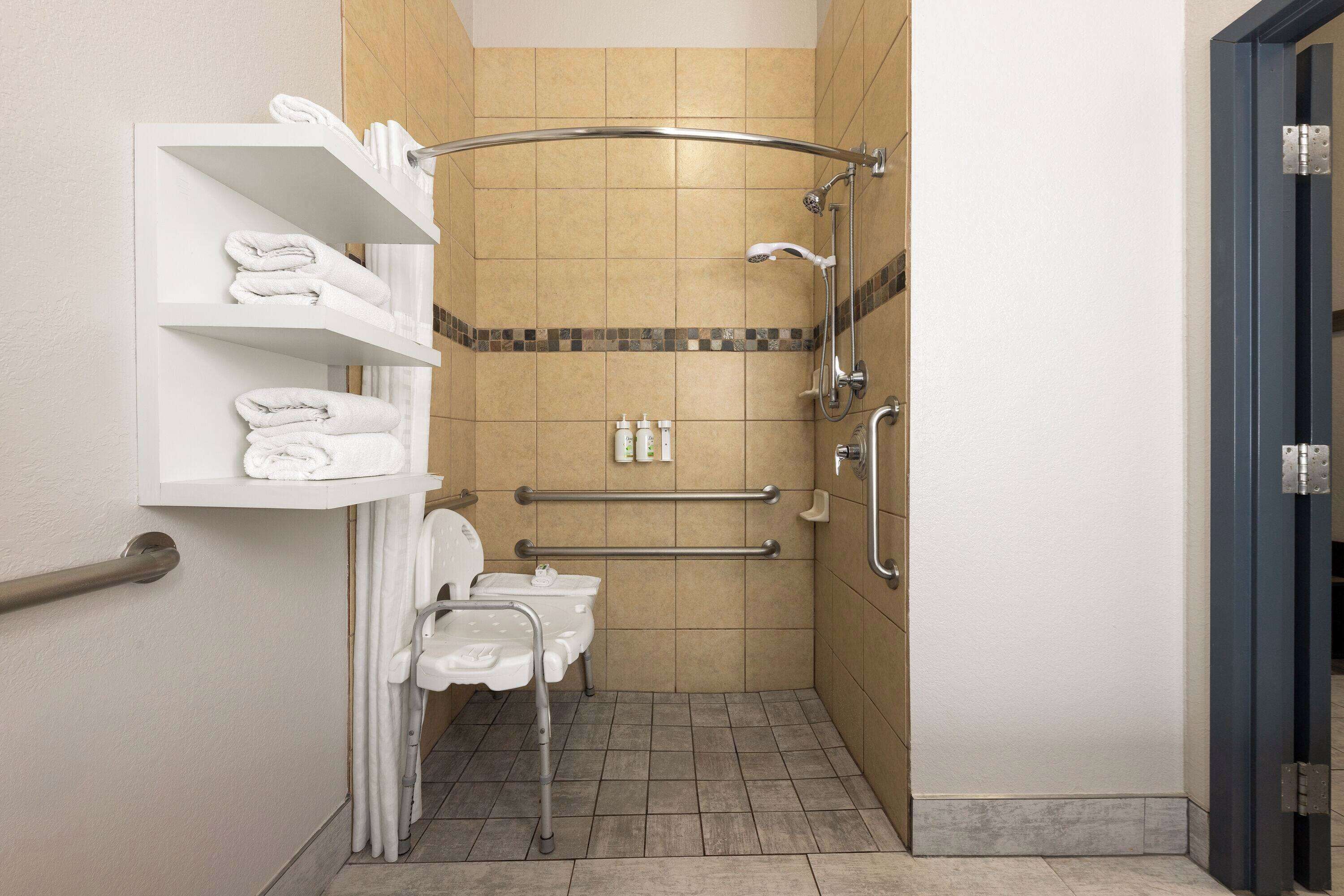 A small, accessible bathroom with a toilet, handrails, a shower, and a shelf with towels.