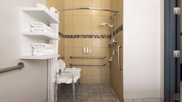 A small, accessible bathroom with a toilet, handrails, a shower, and a shelf with towels.