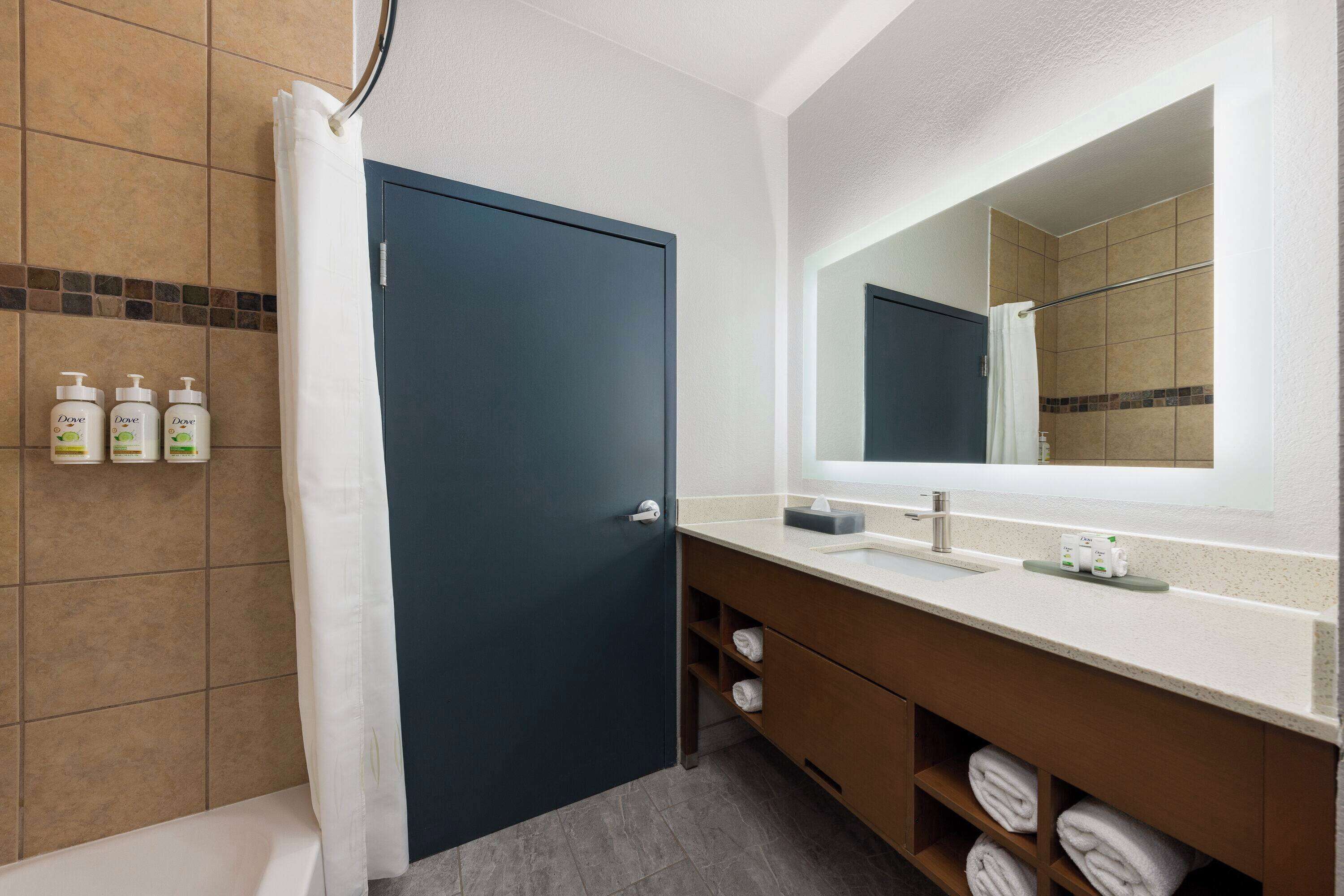 Une salle de bain avec une porte bleu foncé, un lavabo blanc et un miroir au-dessus. On y trouve des articles de toilette accrochés au mur et des serviettes rangées dans un meuble en bois.