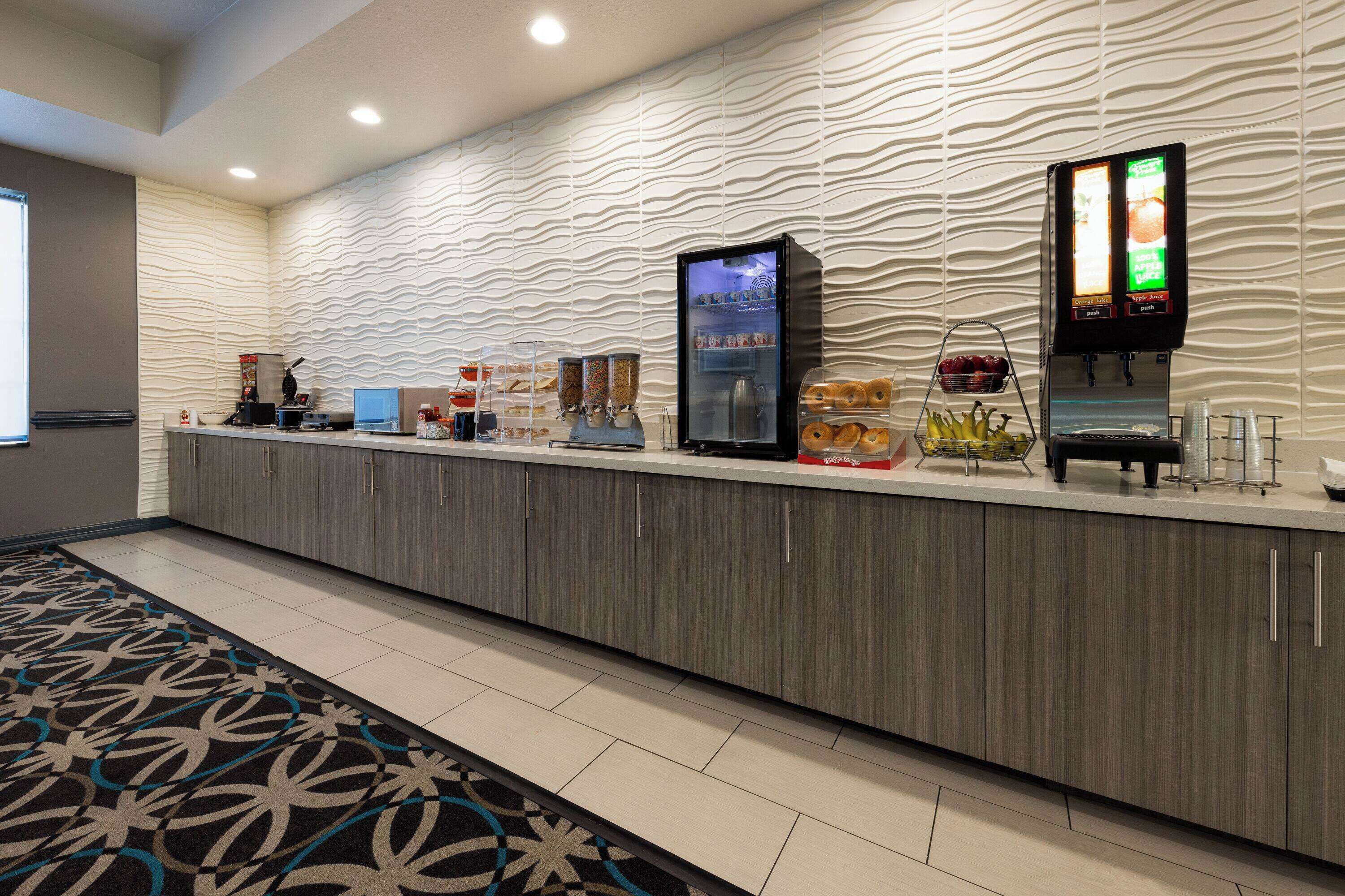 A hotel reception area with a modern design, including a textured wall, a coffee machine, a refrigerated display case, and a self-service beverage station.
