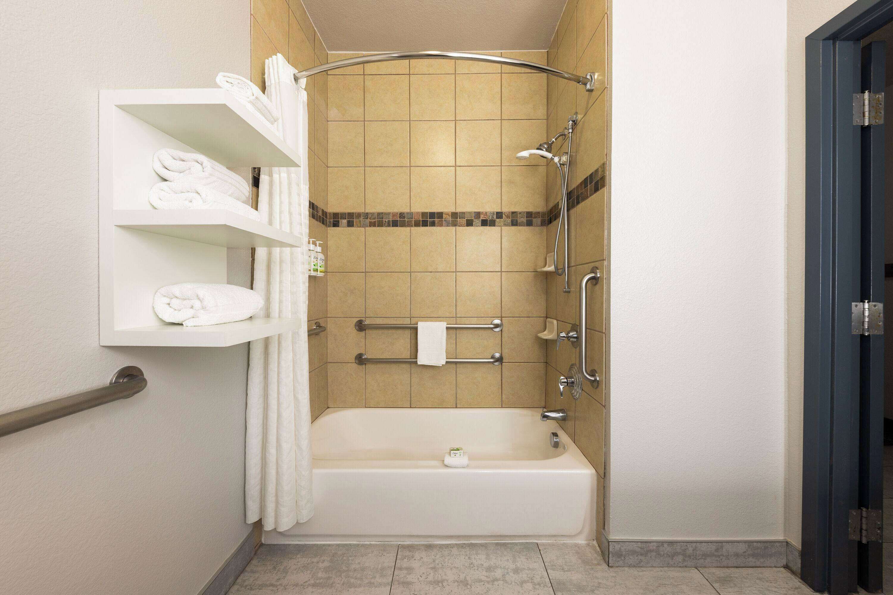 A bathroom with a bathtub, a shelf with towels, a shower, and a handrail.