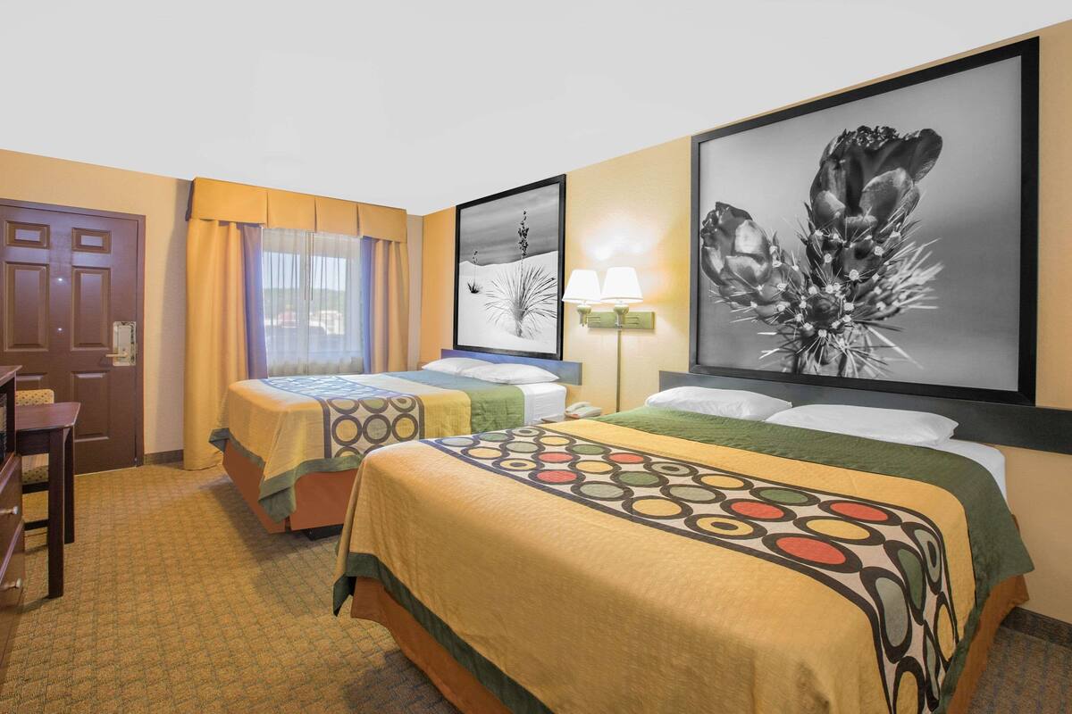 A hotel room with two beds, a desk, a chair, a lamp, and artwork on the walls.