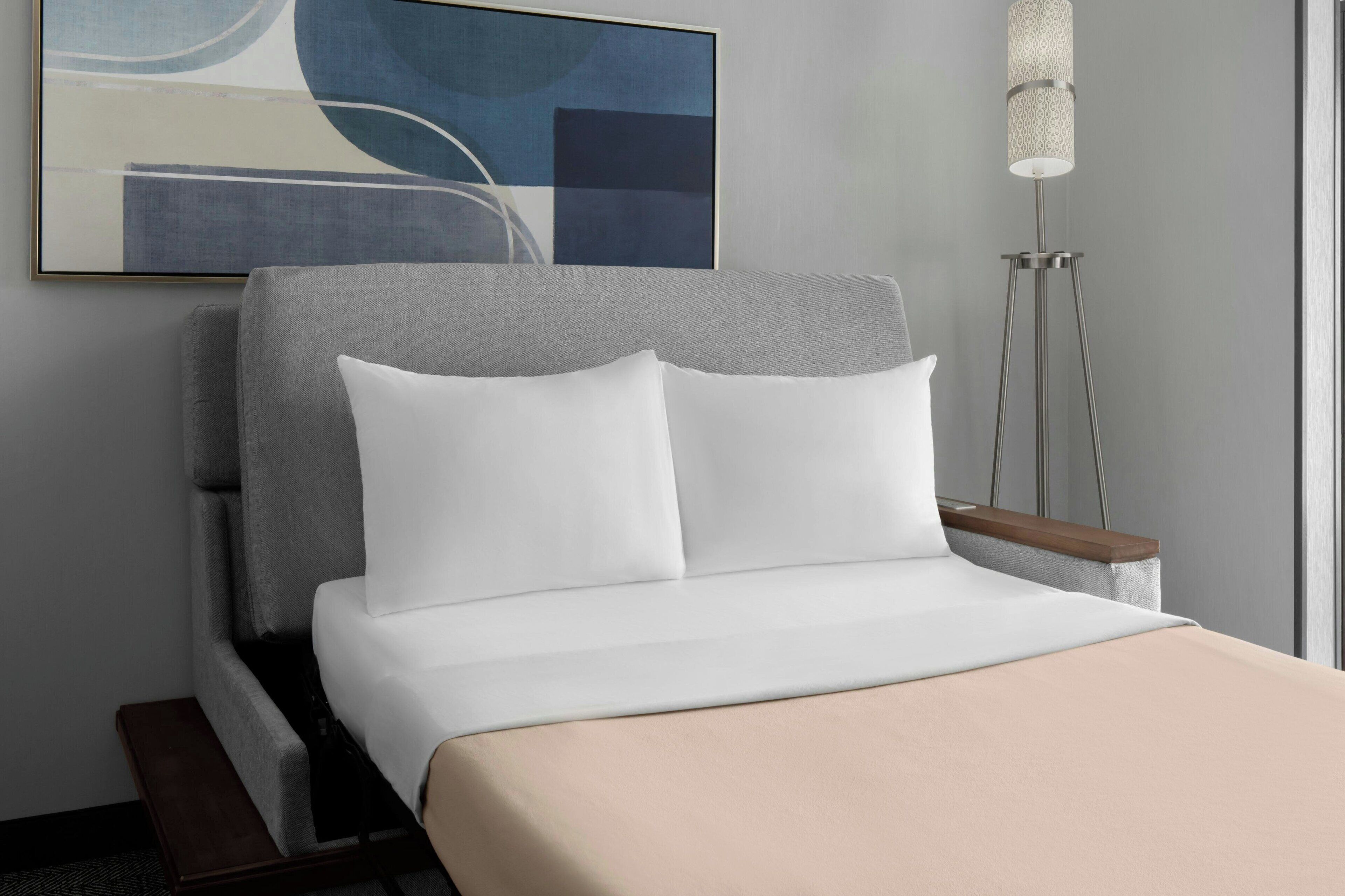 Moderni hotellihuone, jossa on sänky, yöpöytä, lattiavalaisin ja seinällä abstrakti taideteos.