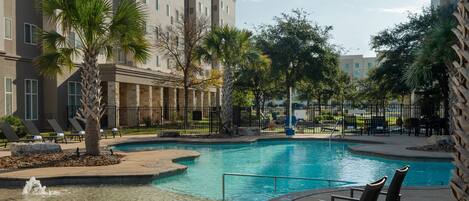 A swimming pool with lounge chairs and palm trees, surrounded by a fence and a building with multiple windows.