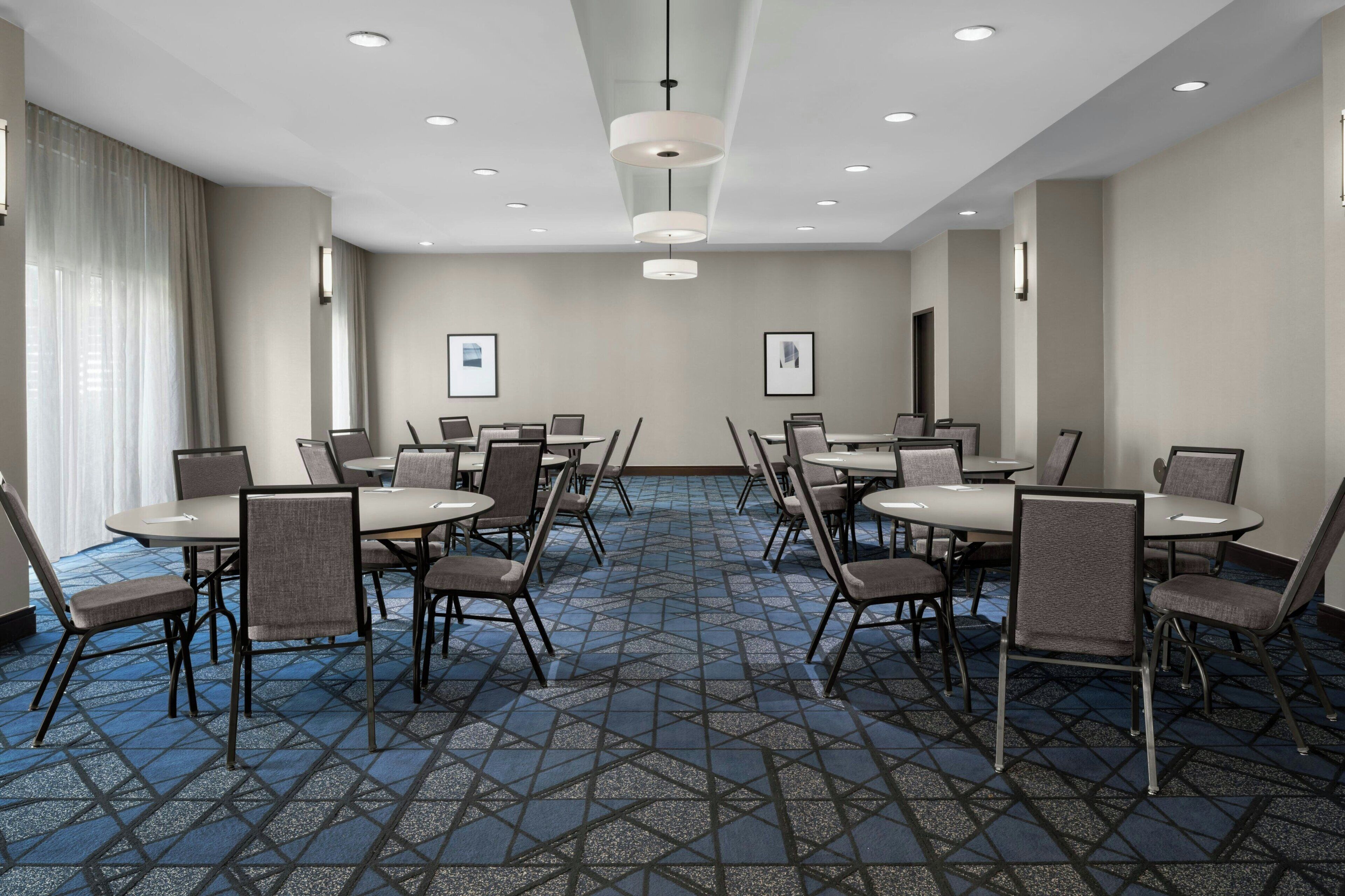 Une salle de conférence dotée de tables rondes et de chaises, d’un tapis à motifs et de luminaires encastrés au plafond.