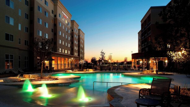 A hotel pool area with multiple fountains, surrounded by lounge chairs and a well-lit evening setting.