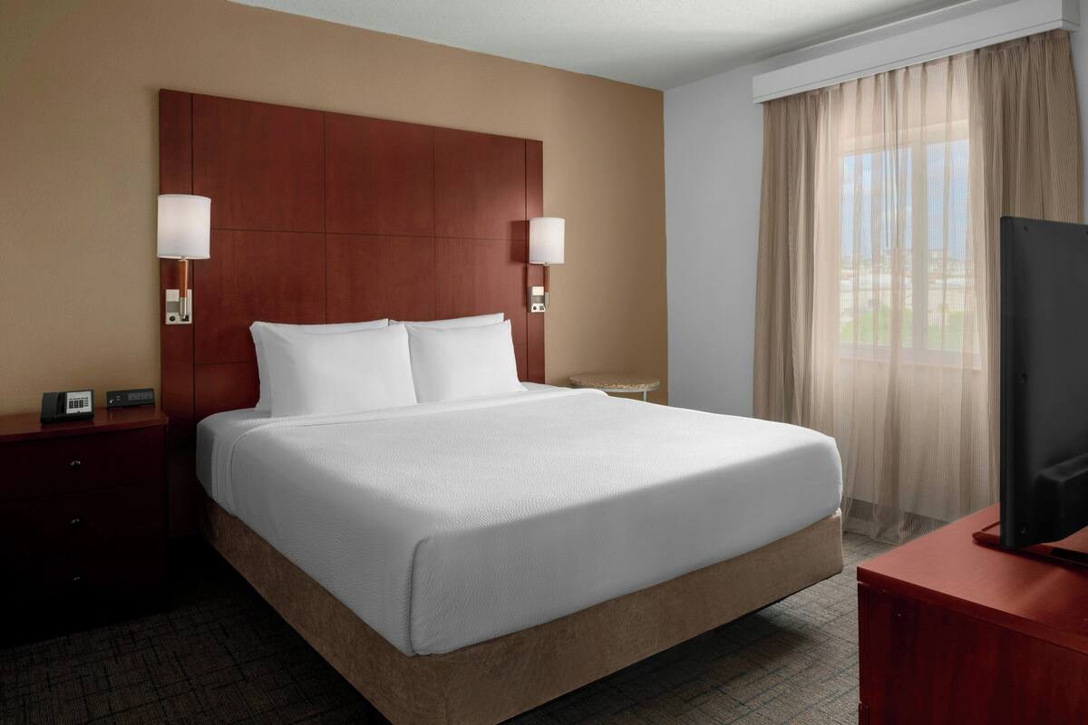 A hotel room with a large bed, two bedside lamps, a TV, a nightstand, and a window with curtains.