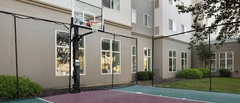 An outdoor basketball court with a net, surrounded by a fence, and adjacent to a building with windows and a door.