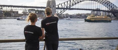 Two people in matching black t-shirts with "HOME BACKPACKERS" on the back, standing by a railing overlooking a body of water with a large bridge in the background.