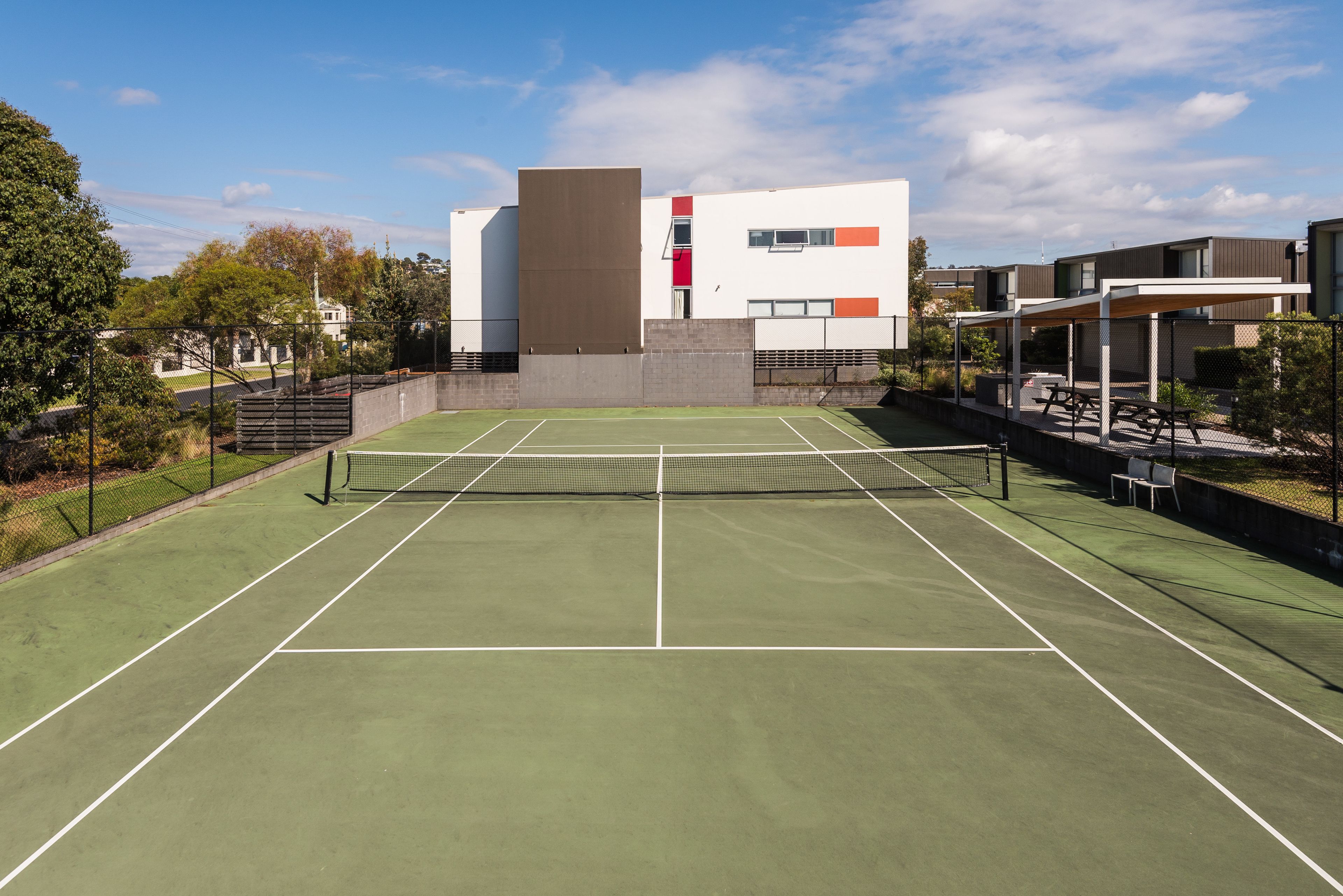 Tenniskenttä, jossa on verkko, ja kentän ympärillä on aita. Taustalla näkyy rakennuksia.