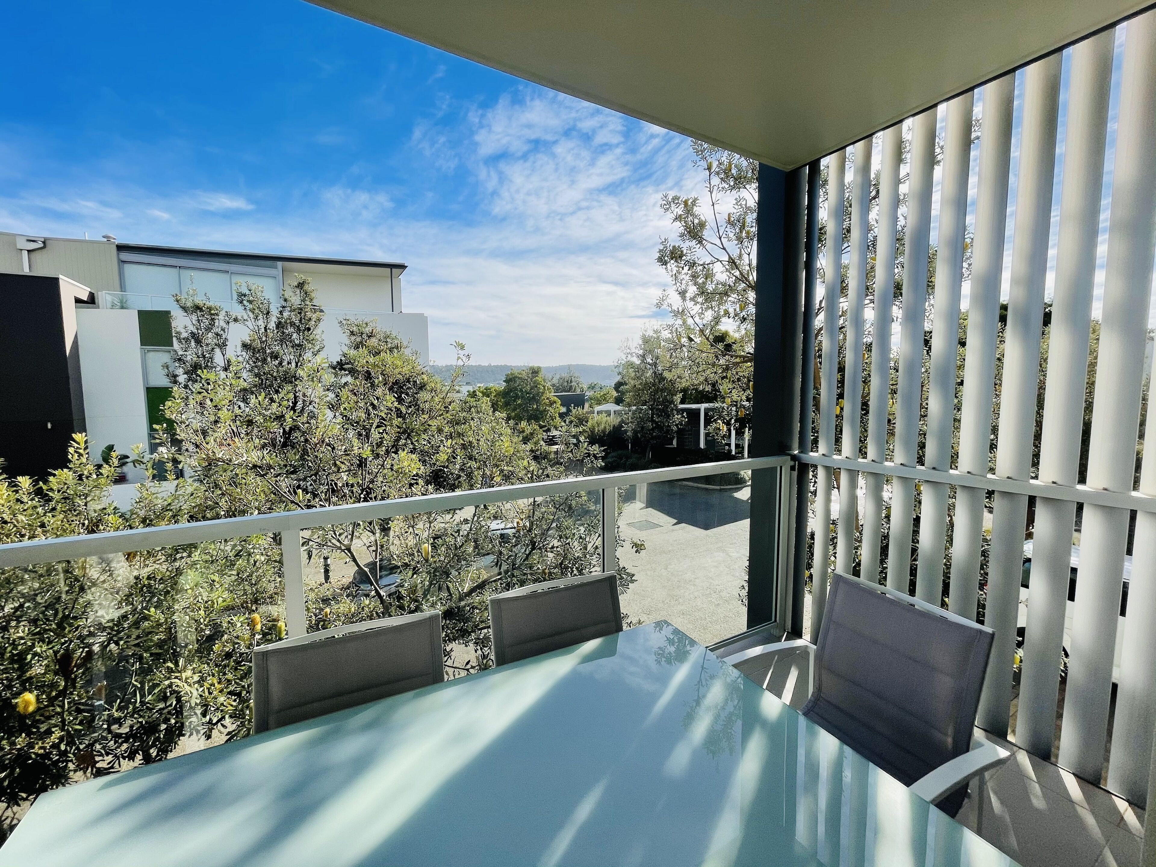 A balcony with a glass table and chairs, overlooking a garden and modern buildings.