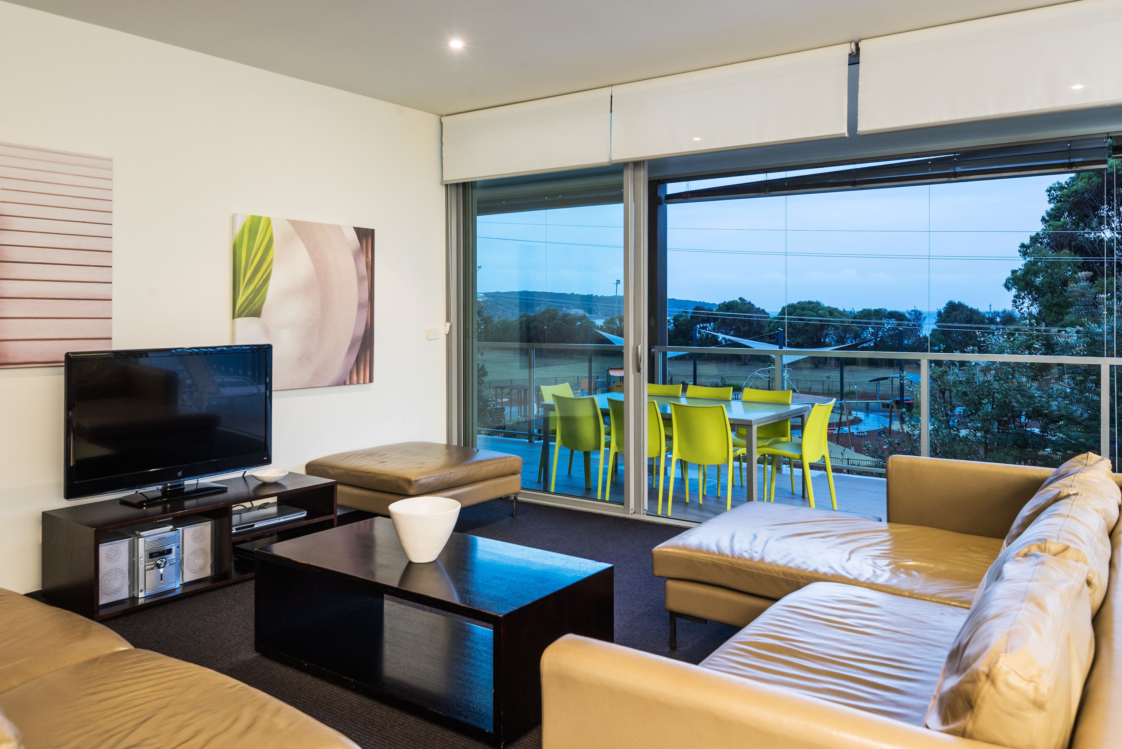A modern living room with a flat-screen TV, comfortable seating, a coffee table, and large windows offering a view of the outdoors.