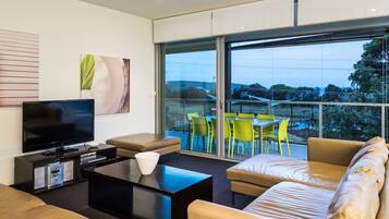 A modern living room with a flat-screen TV, comfortable seating, a coffee table, and large windows offering a view of the outdoors.