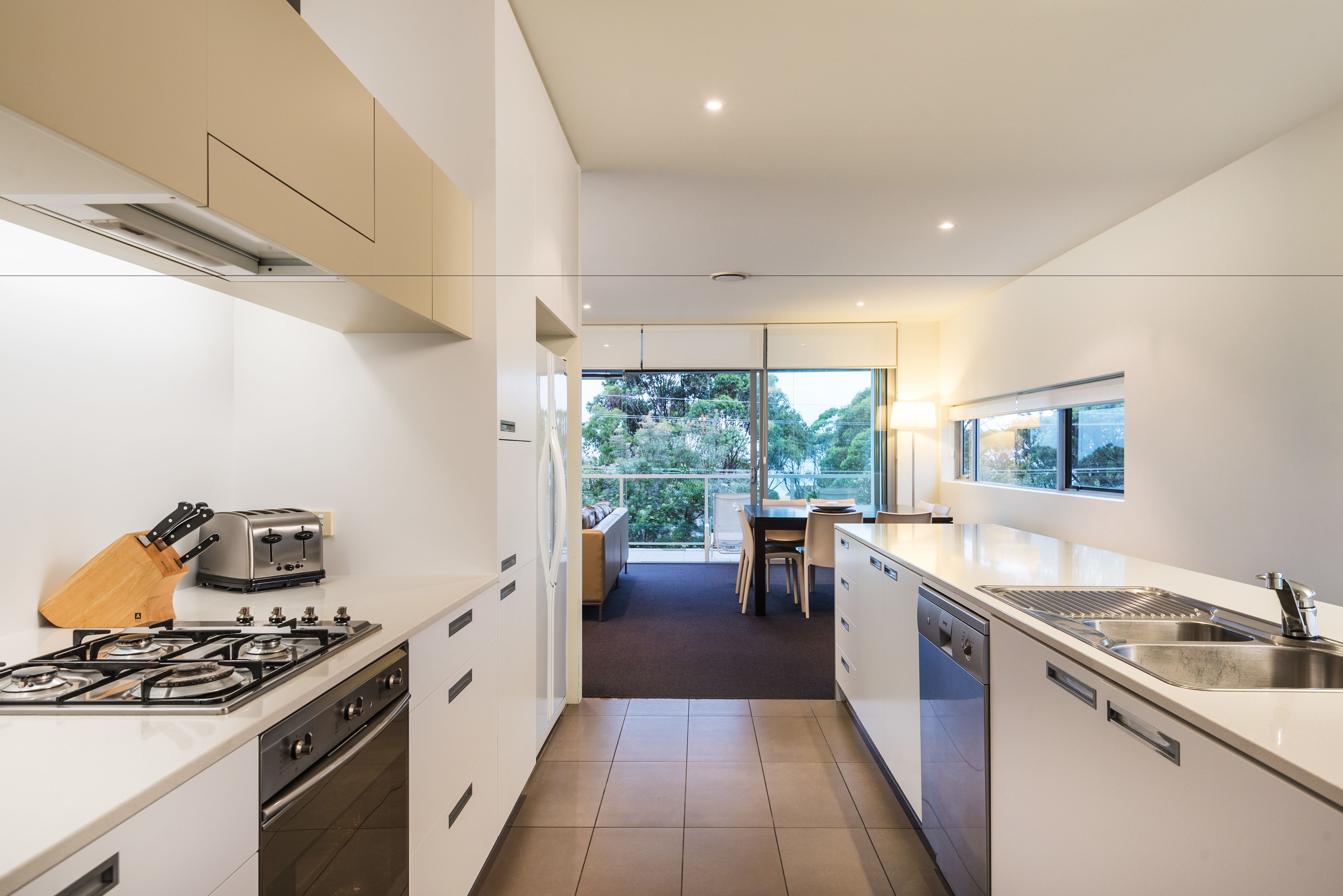 Una cocina moderna con estufa a gas, tostadora y lavavajillas. Se ve un área de comedor con mesa y sillas a través de un vidrio.