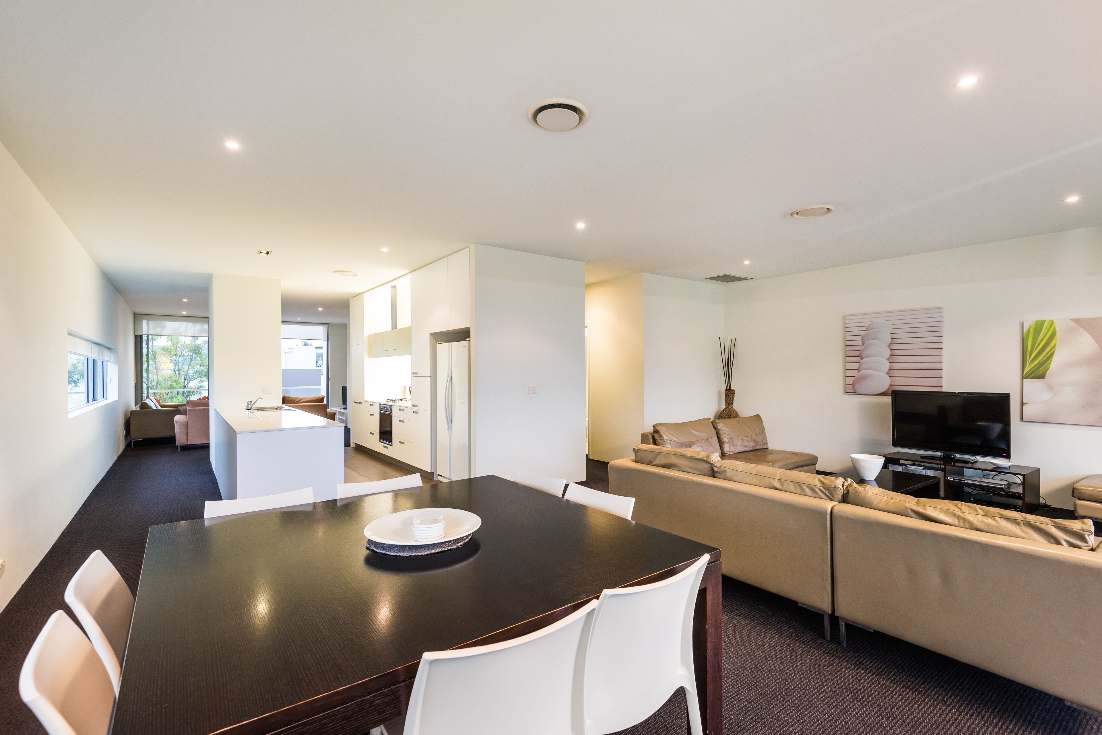 A modern living room with a dark wooden dining table, white chairs, a beige sofa, and a flat-screen TV.