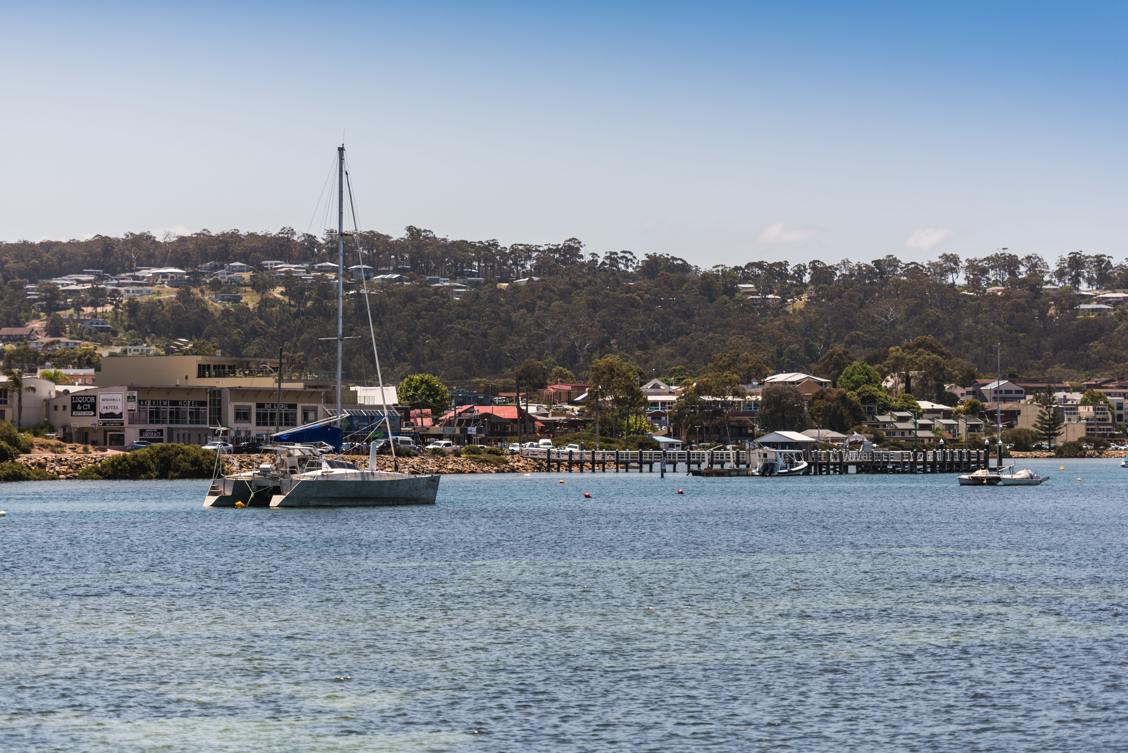A waterfront view with a sailboat docked near a pier, surrounded by residential buildings and a hilly landscape in the background.