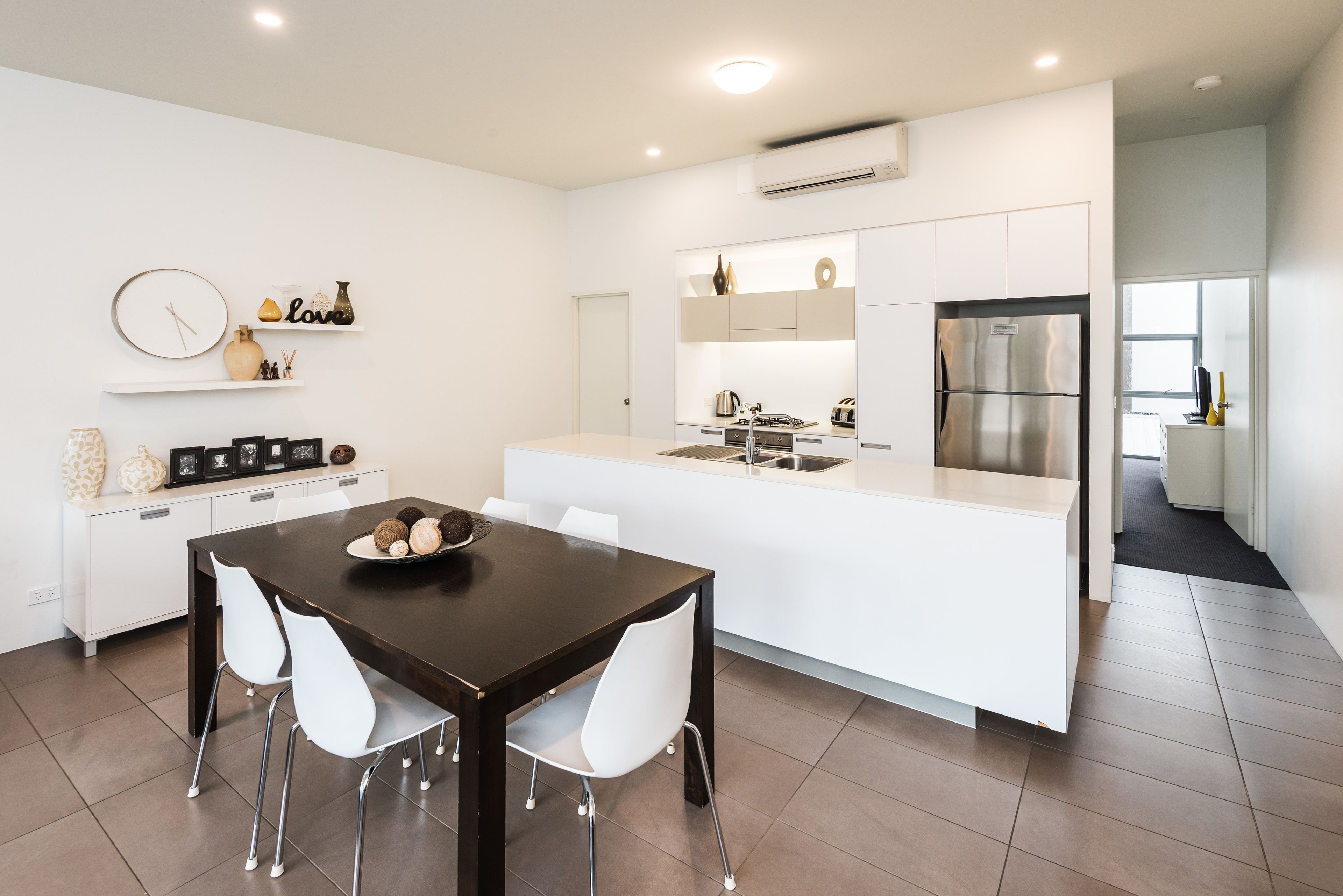 Cocina moderna con armarios blancos, una isla central y un comedor con una mesa oscura y sillas blancas.