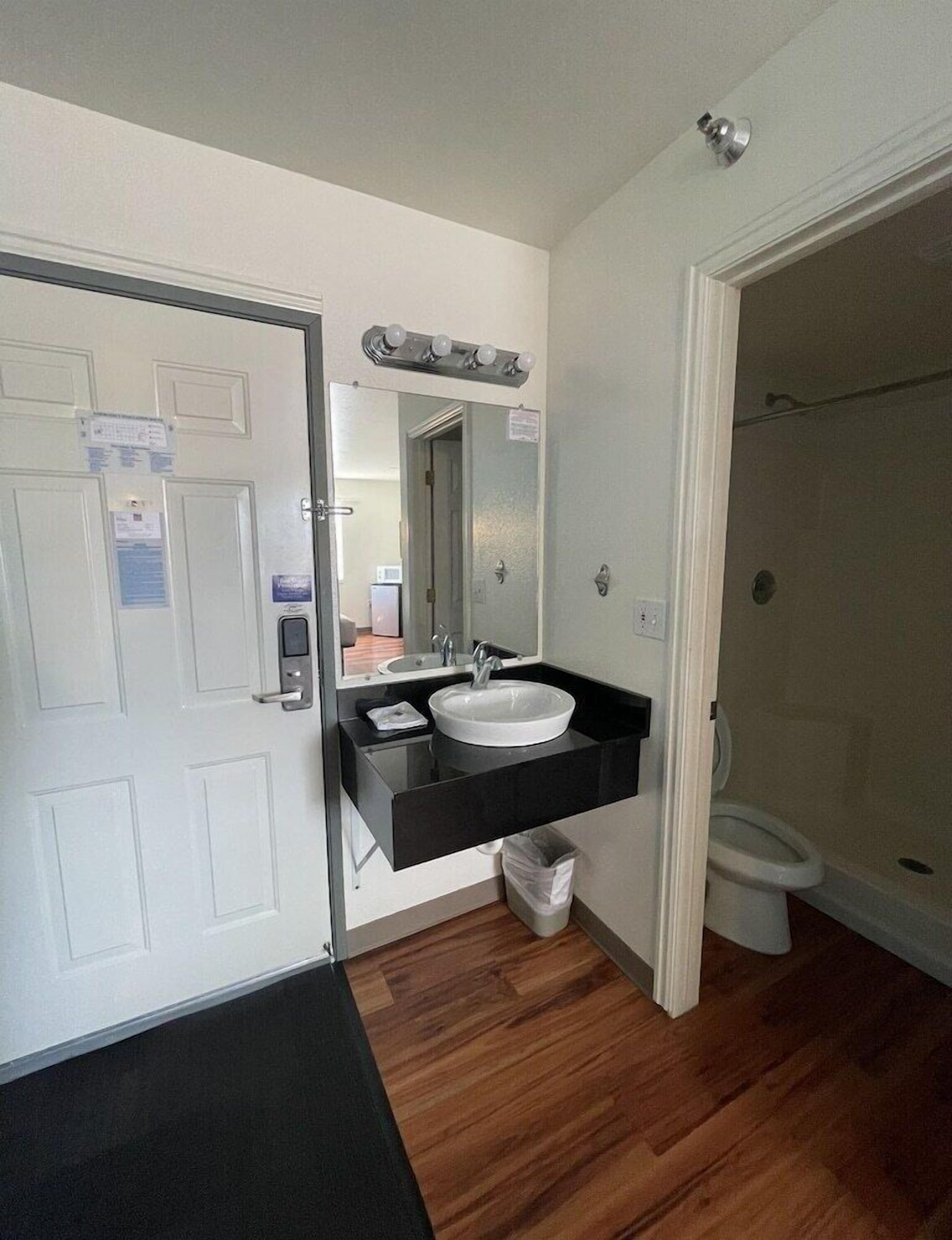 A bathroom with a white sink, black countertop, and a mirror above the sink. There is a white toilet and a shower area visible.