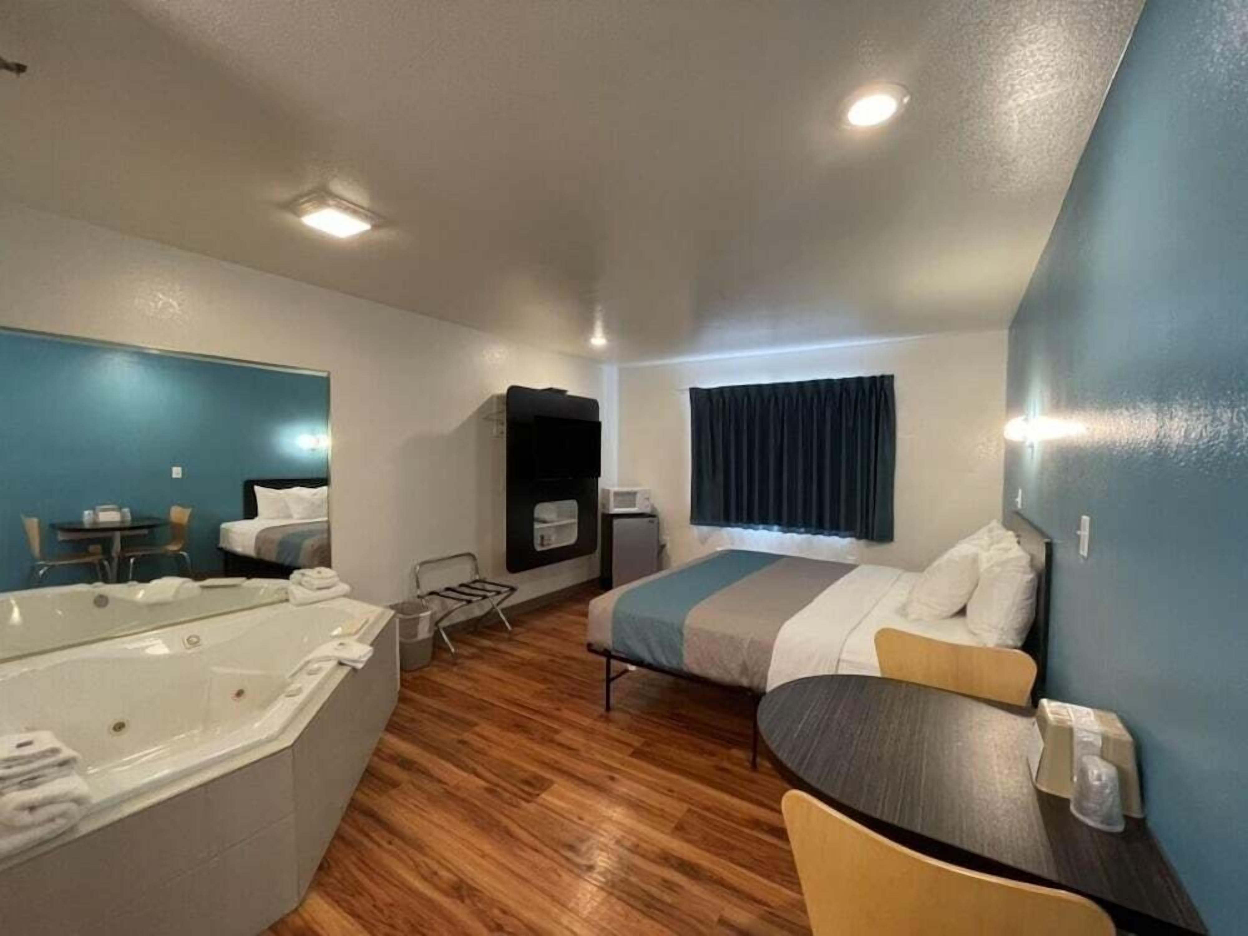 A compact hotel room with a bed, a small table, a chair, a sofa, a bathtub, and a microwave.