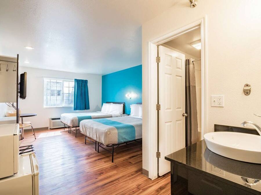 A hotel room with two beds, a large window with curtains, a bathroom with a sink and mirror, and a kitchenette area.