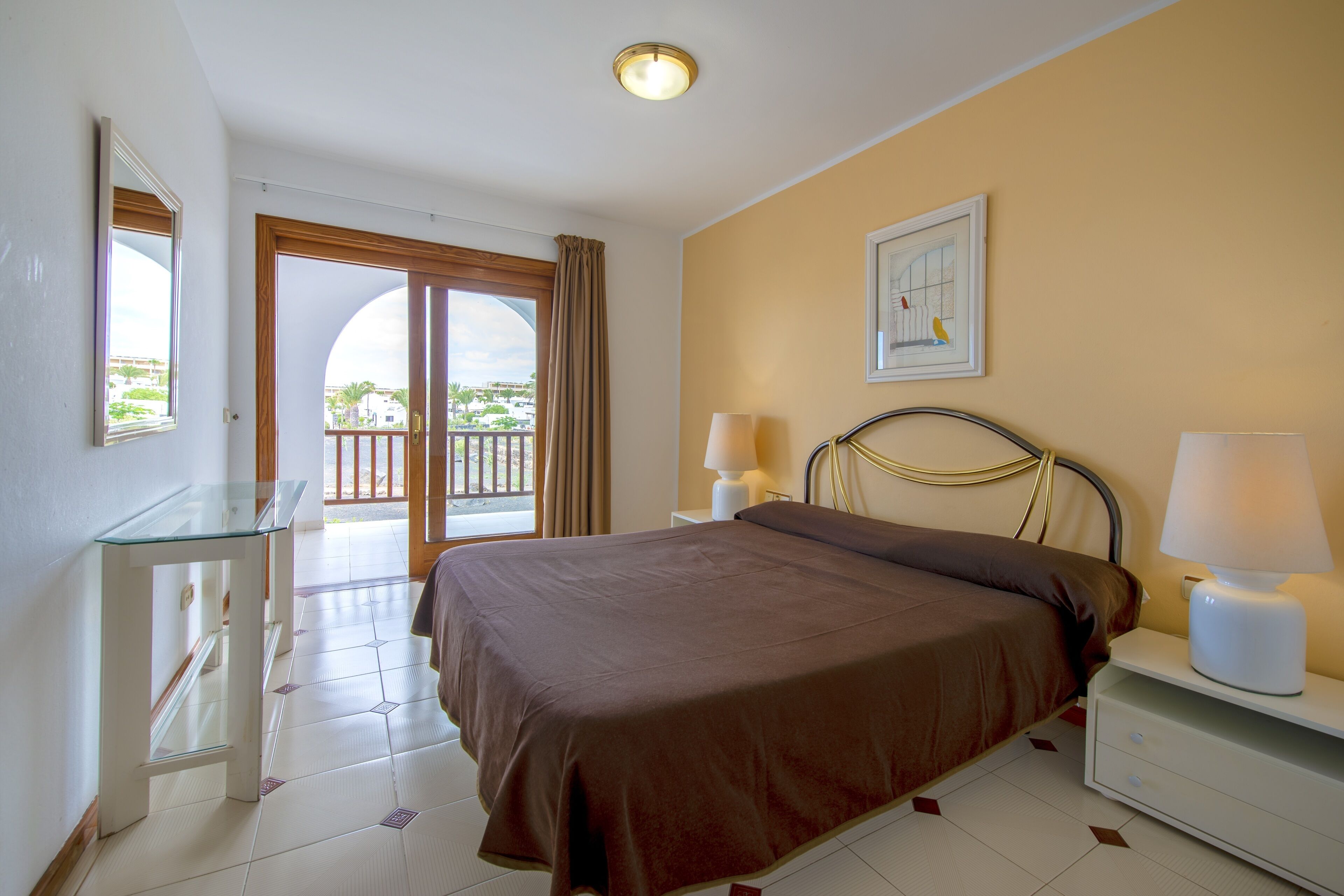 A hotel room with a bed, bedside lamps, a glass table, a door leading to a balcony, and a framed picture on the wall.