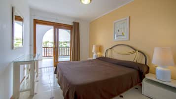 A hotel room with a bed, bedside lamps, a glass table, a door leading to a balcony, and a framed picture on the wall.