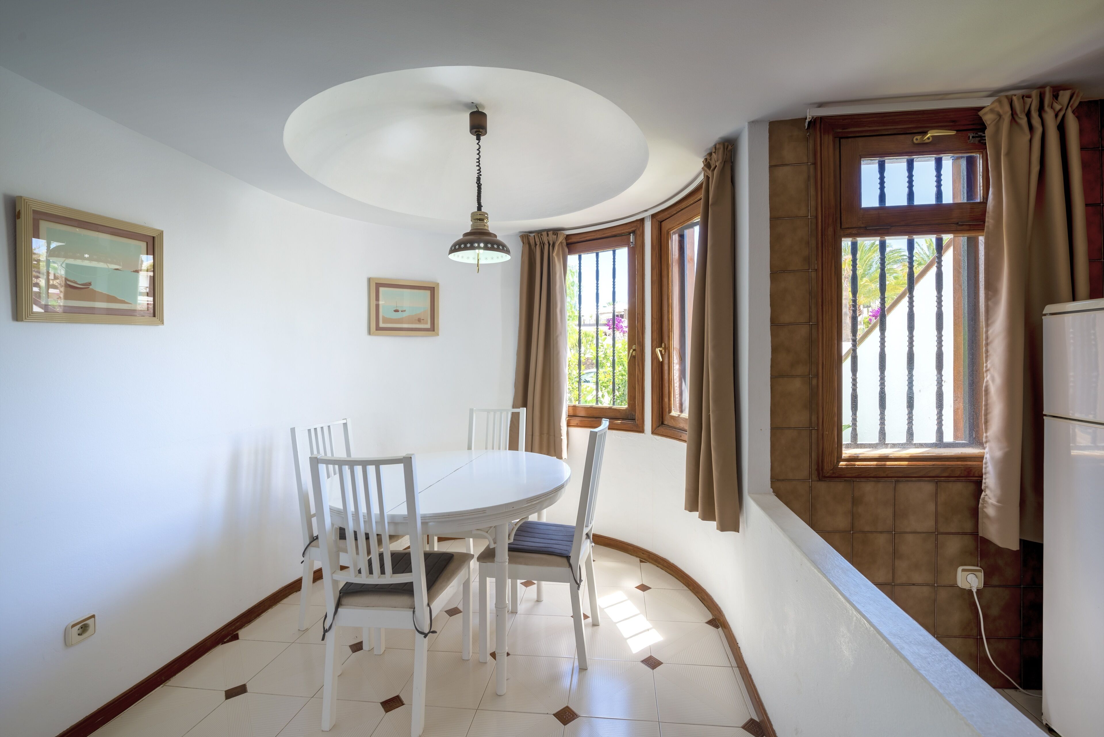 A dining area with a white table and chairs, a hanging light fixture, and a window with curtains.