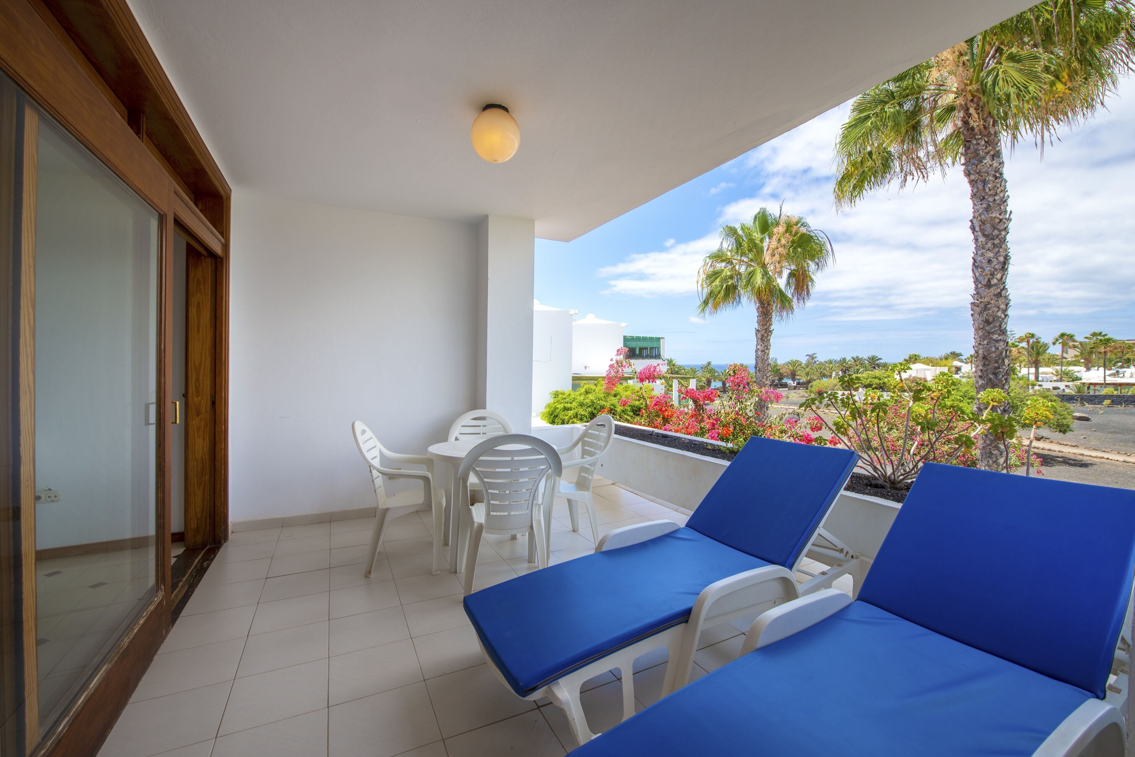A balcony with white chairs and blue cushions, overlooking a garden with palm trees and flowers.