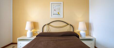 A bedroom with a bed, two bedside tables with lamps, a framed picture on the wall, and a plain yellow wall.