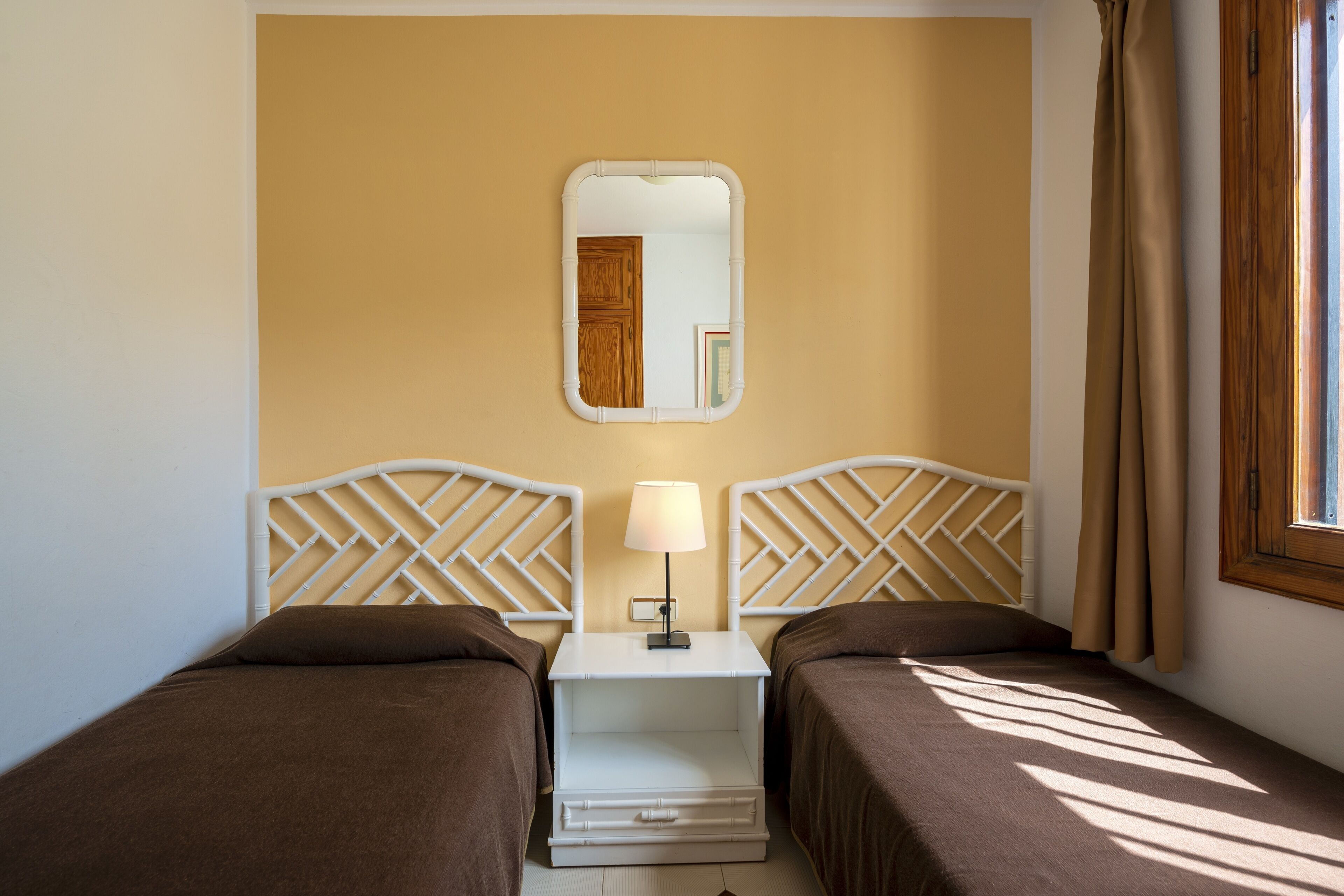 Habitación de hotel con dos camas, una mesita de noche con lámpara, un espejo y una ventana con cortinas.