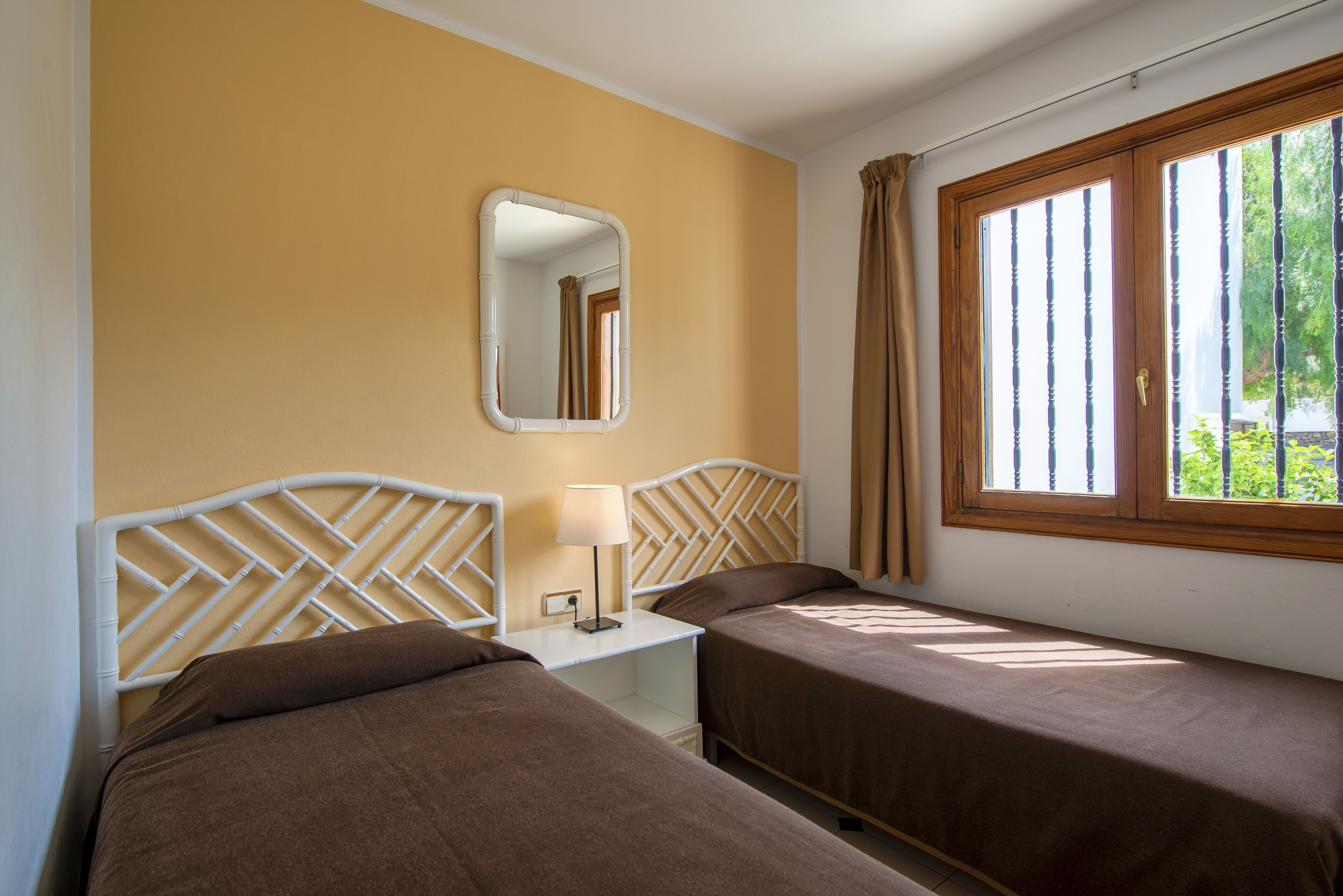 A hotel room with two single beds, a nightstand with a lamp, a mirror, and a window with curtains.