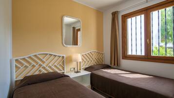 A hotel room with two single beds, a nightstand with a lamp, a mirror, and a window with curtains.