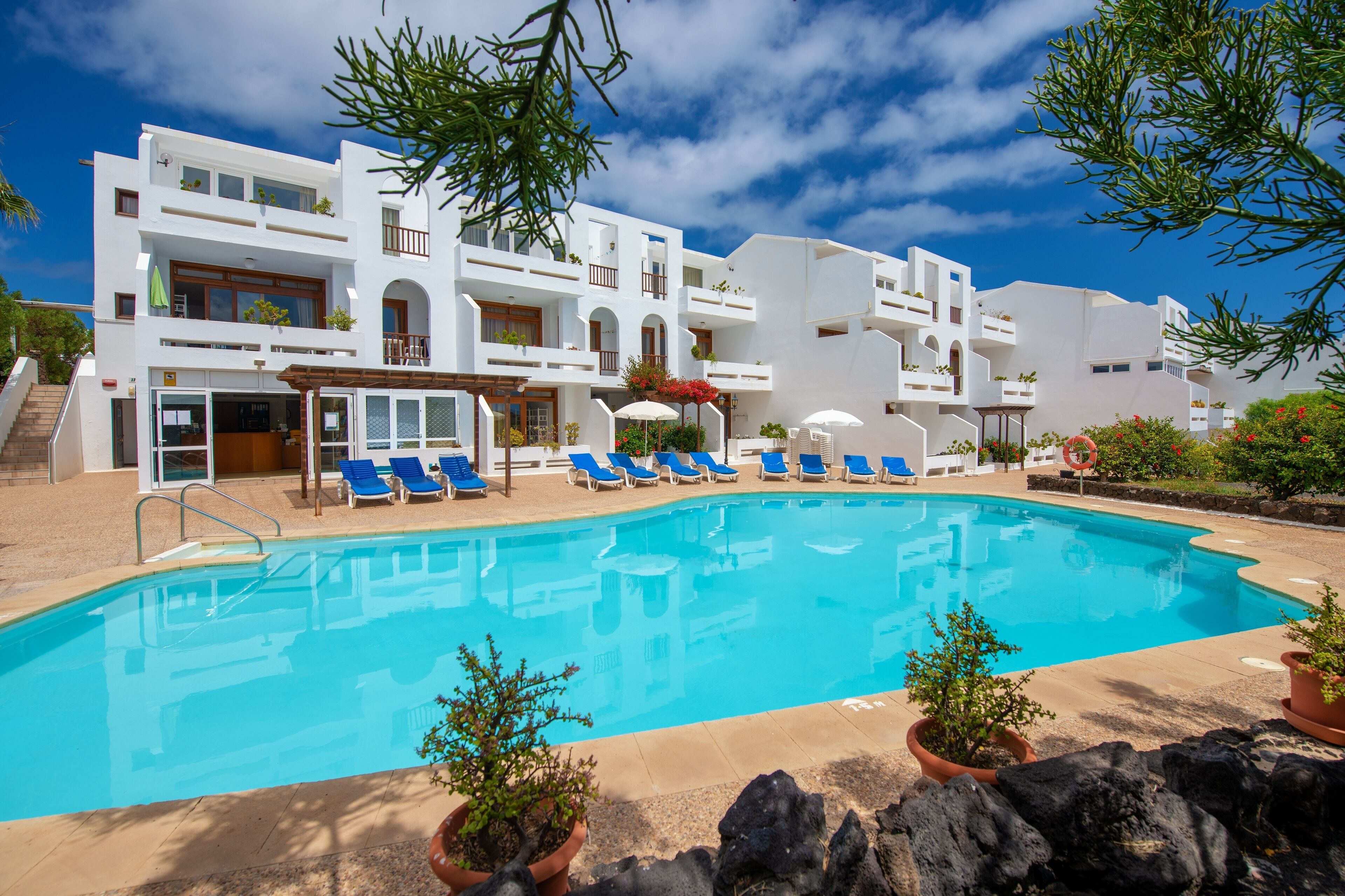 A poolside area with a clear blue pool, white buildings, and blue lounge chairs.