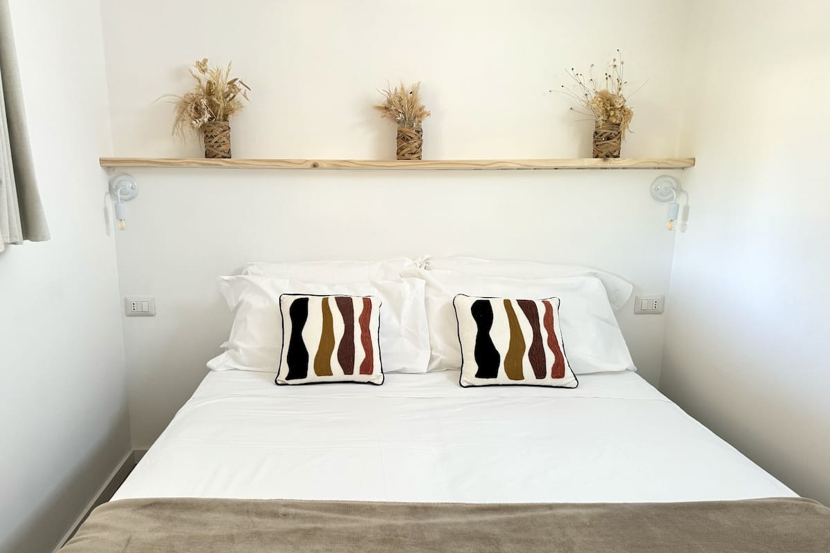 A neatly made bed with white linens, two decorative pillows with a striped pattern, and a brown throw blanket. Above the bed, there are three small potted plants on a wooden shelf.