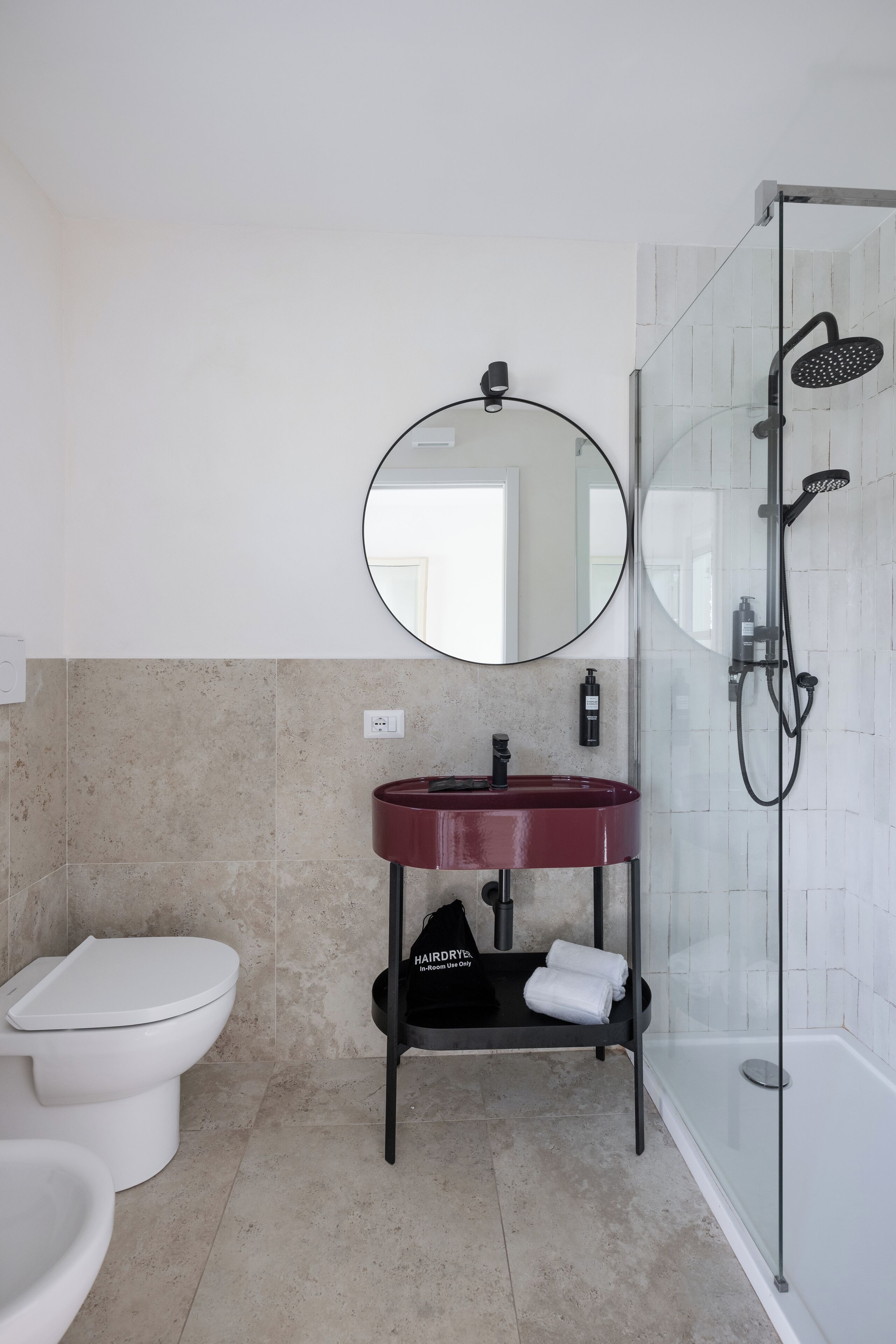 Ein modernes Badezimmer mit Toilette, rundem Spiegel über einem roten Waschbecken und einer Duschkabine.