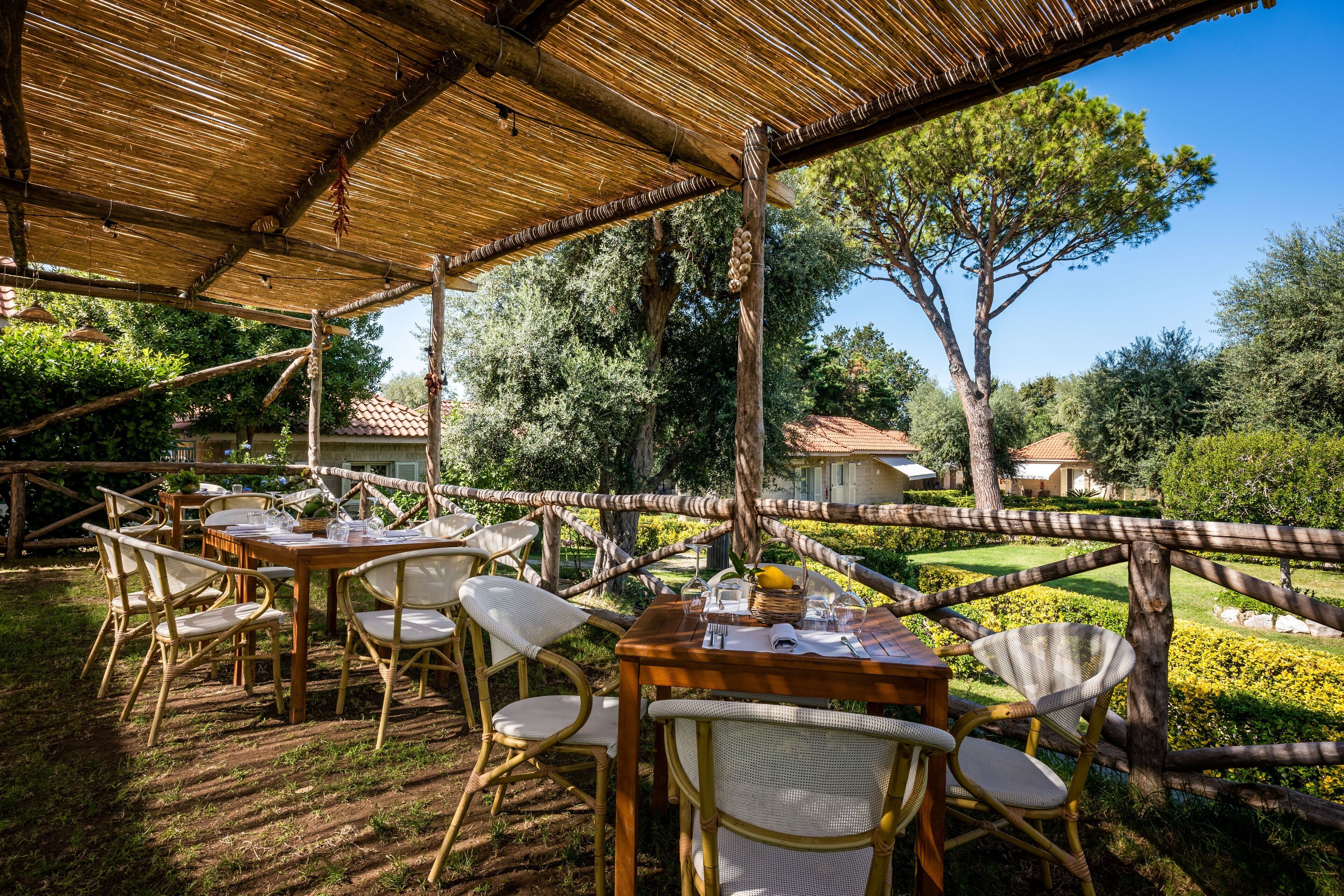 An outdoor dining area with wooden tables and wicker chairs, surrounded by a wooden fence and lush greenery.