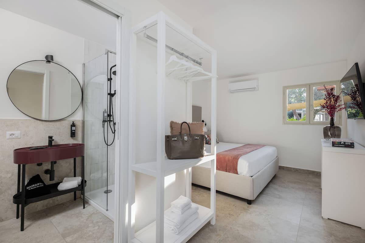 A modern bathroom with a glass shower enclosure, a round mirror, a vanity with a red sink, and a white shelf with towels and a handbag.