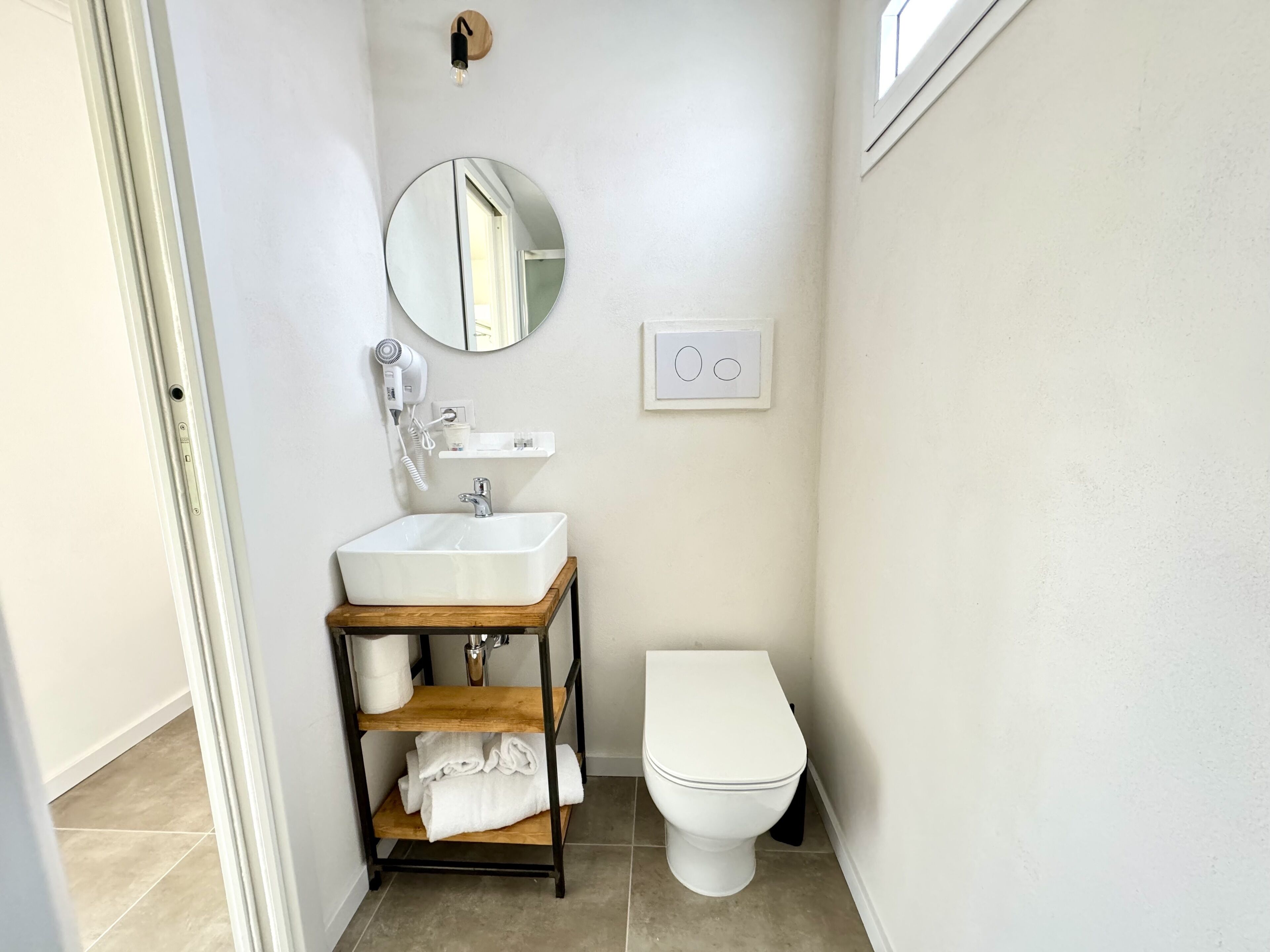 A bathroom with a white sink, a round mirror, a hairdryer, a toilet, and a shelf with towels.
