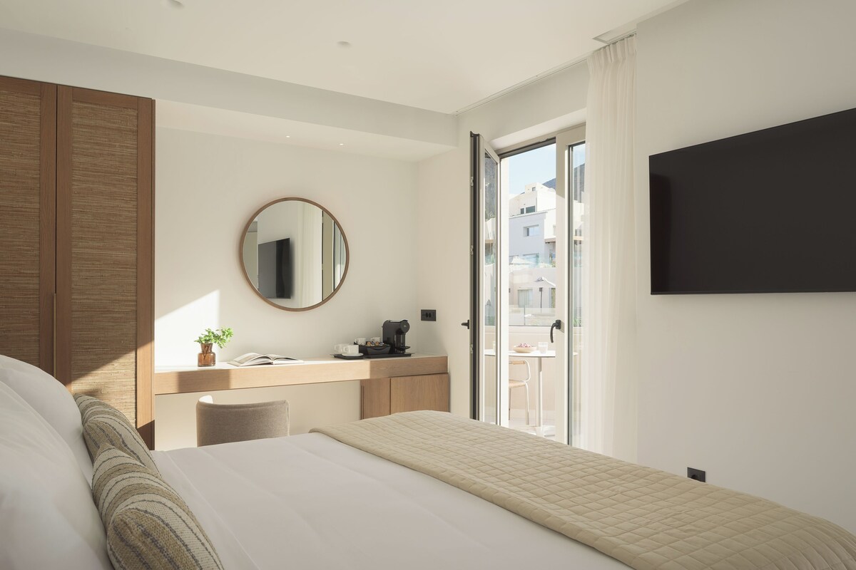 Una habitación de hotel moderna con cama, escritorio con cafetera, espejo redondo y televisor en la pared.