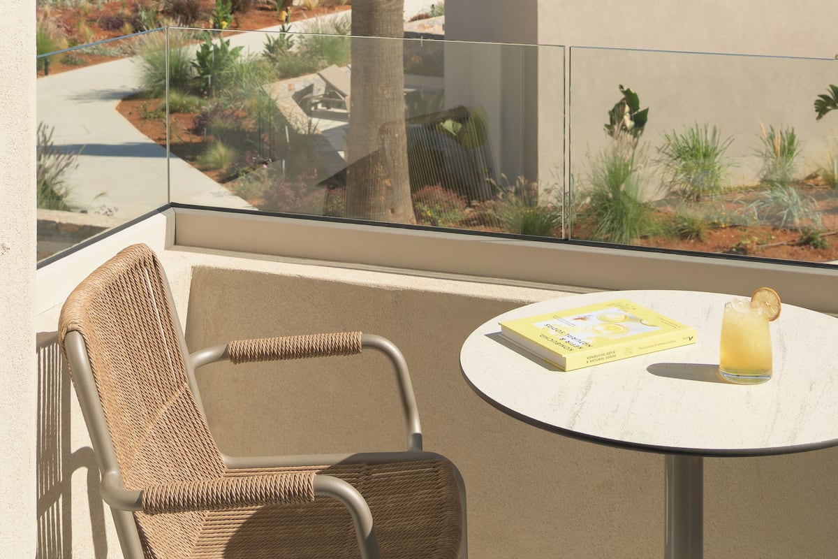 A balcony with a wicker chair, a small table with a book and a drink, and a view of a palm tree and buildings.