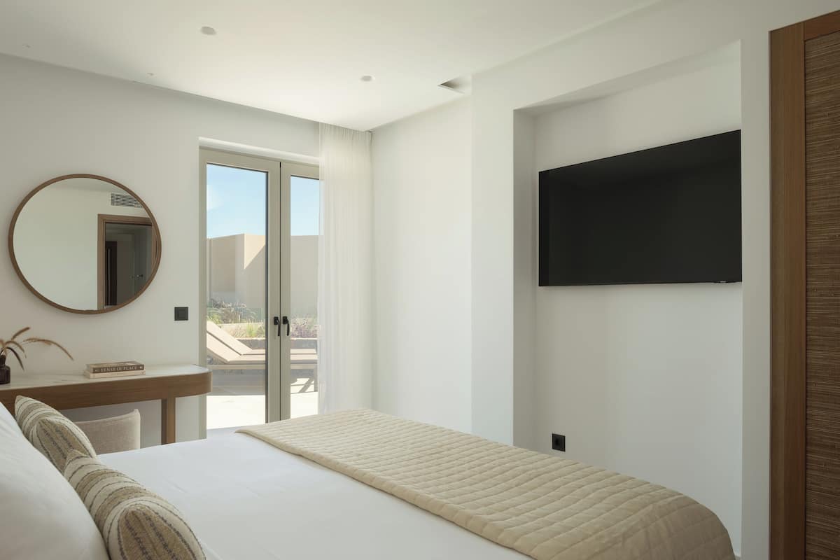 A bedroom with a bed, a desk, a mirror, a TV, and a view of the outdoors.